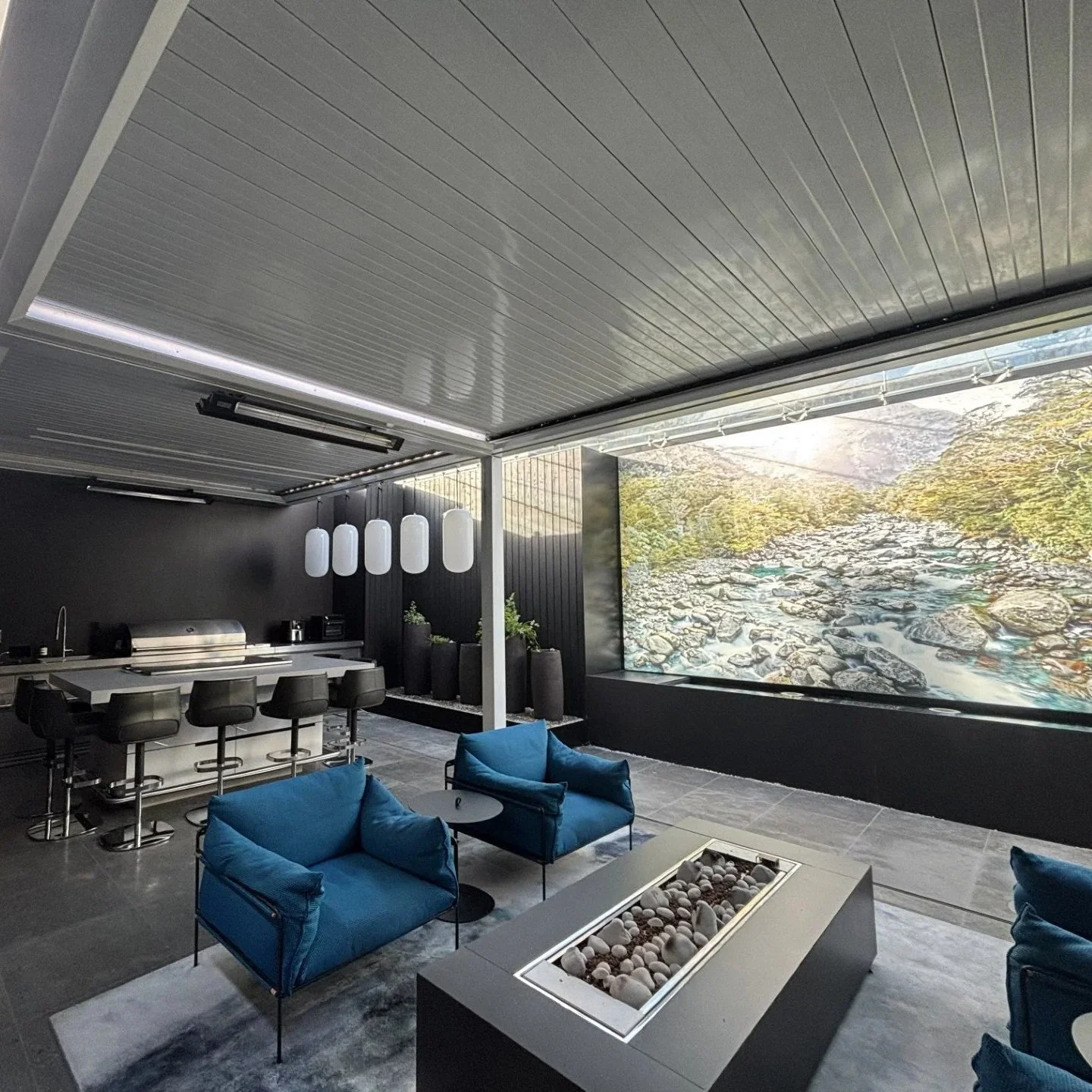 Modern indoor lounge area with blue armchairs, a fire pit table, a high-top breakfast bar with black stools, hanging pendant lights, and a large landscape mural of a river scene.