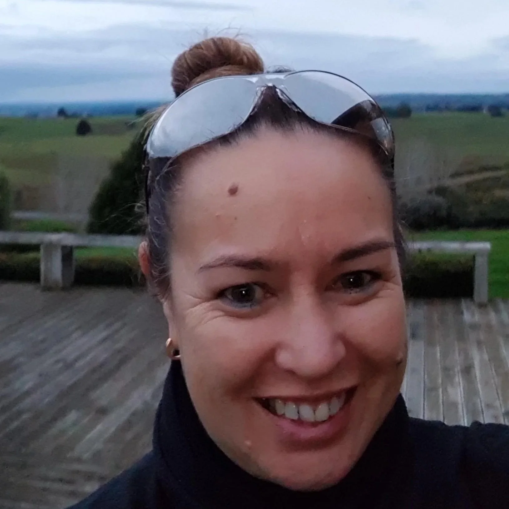 Close-up of a smiling woman with sunglasses on her head outdoors, with a green field and cloudy sky in the background.