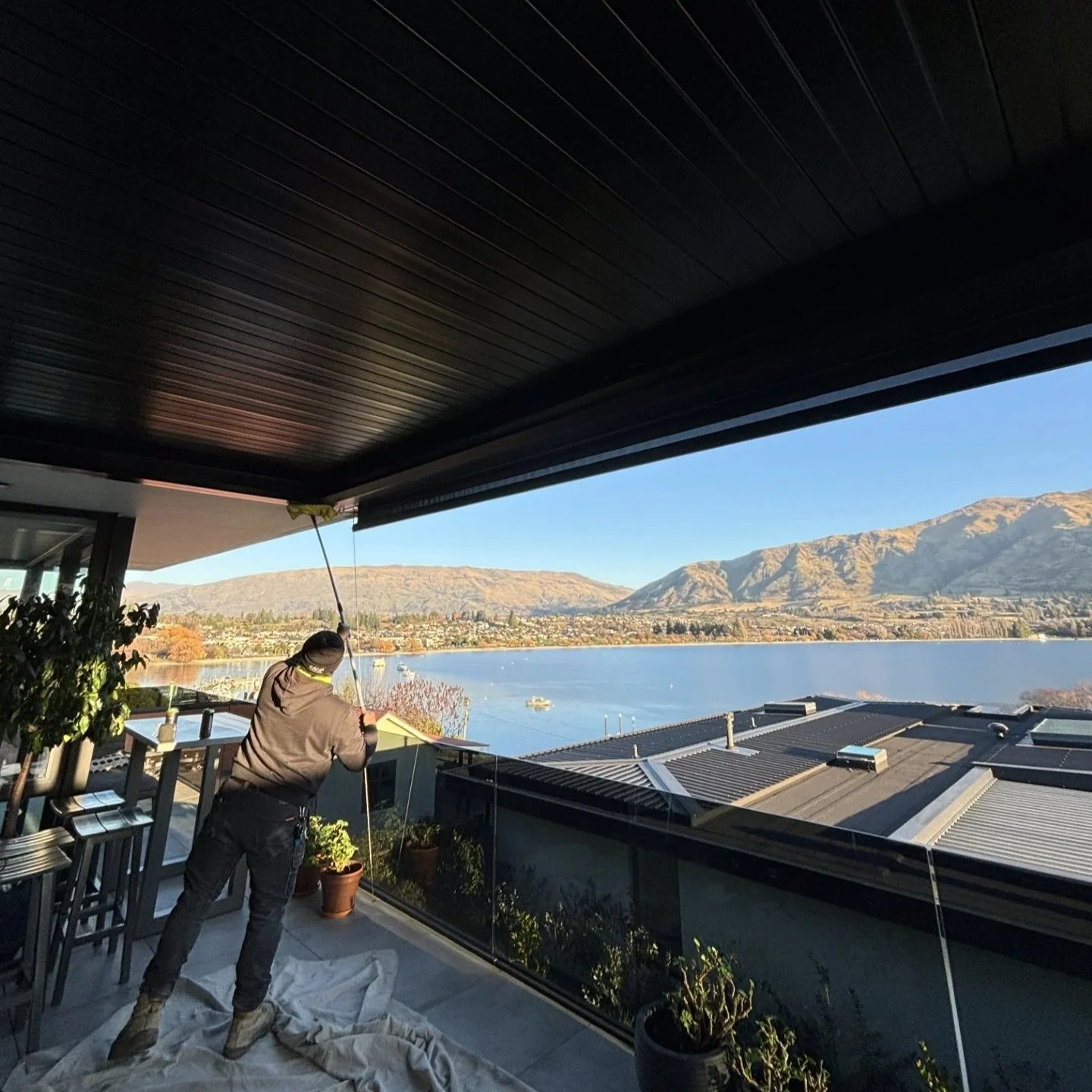 Person cleaning or inspecting the awning from a balcony overlooking a lake and mountain landscape under a clear blue sky.