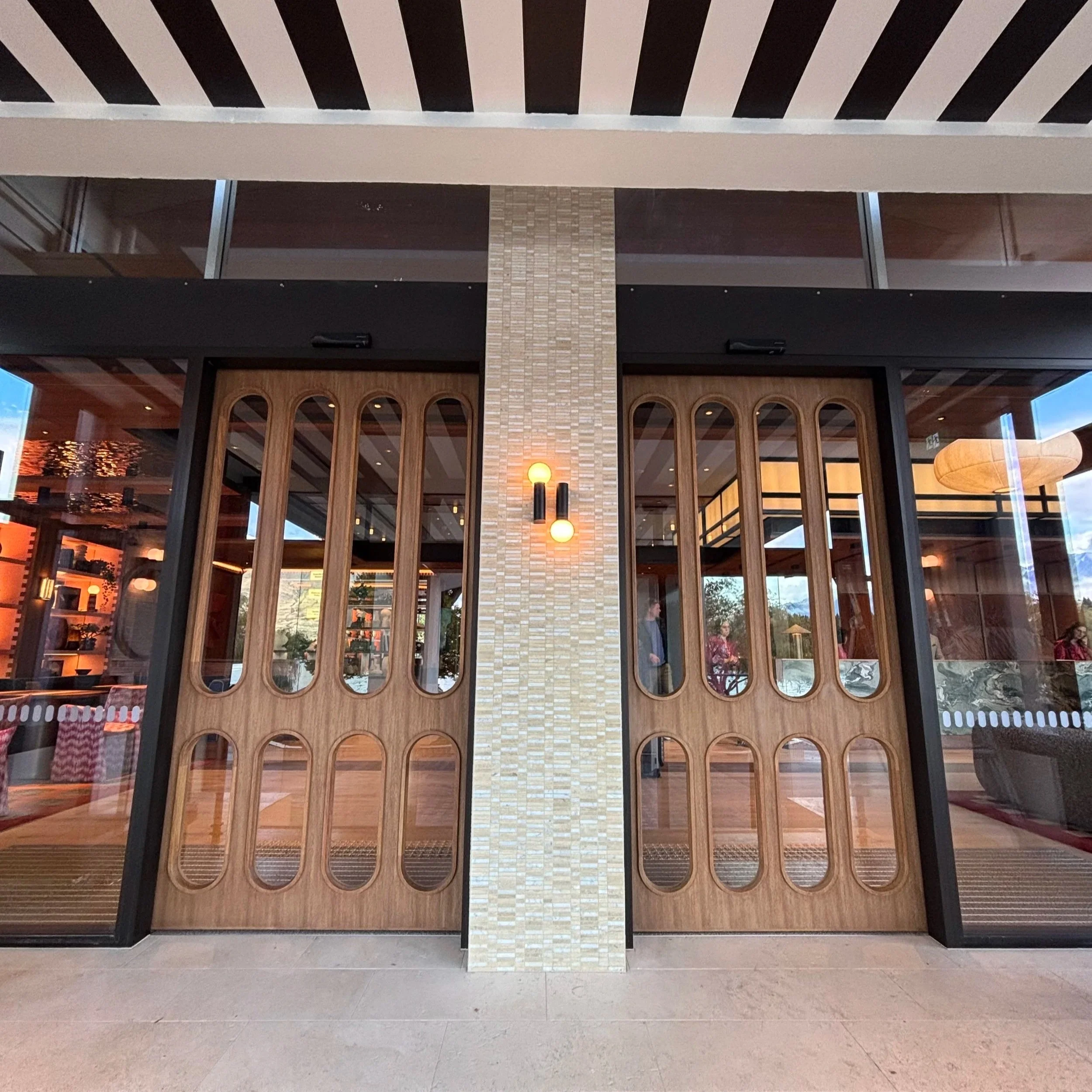 The image shows a modern restaurant entrance with large glass windows and doors. There is wood paneling above the door and glass display shelves inside. Two hanging light fixtures are visible through the glass, along with some people and decor inside.