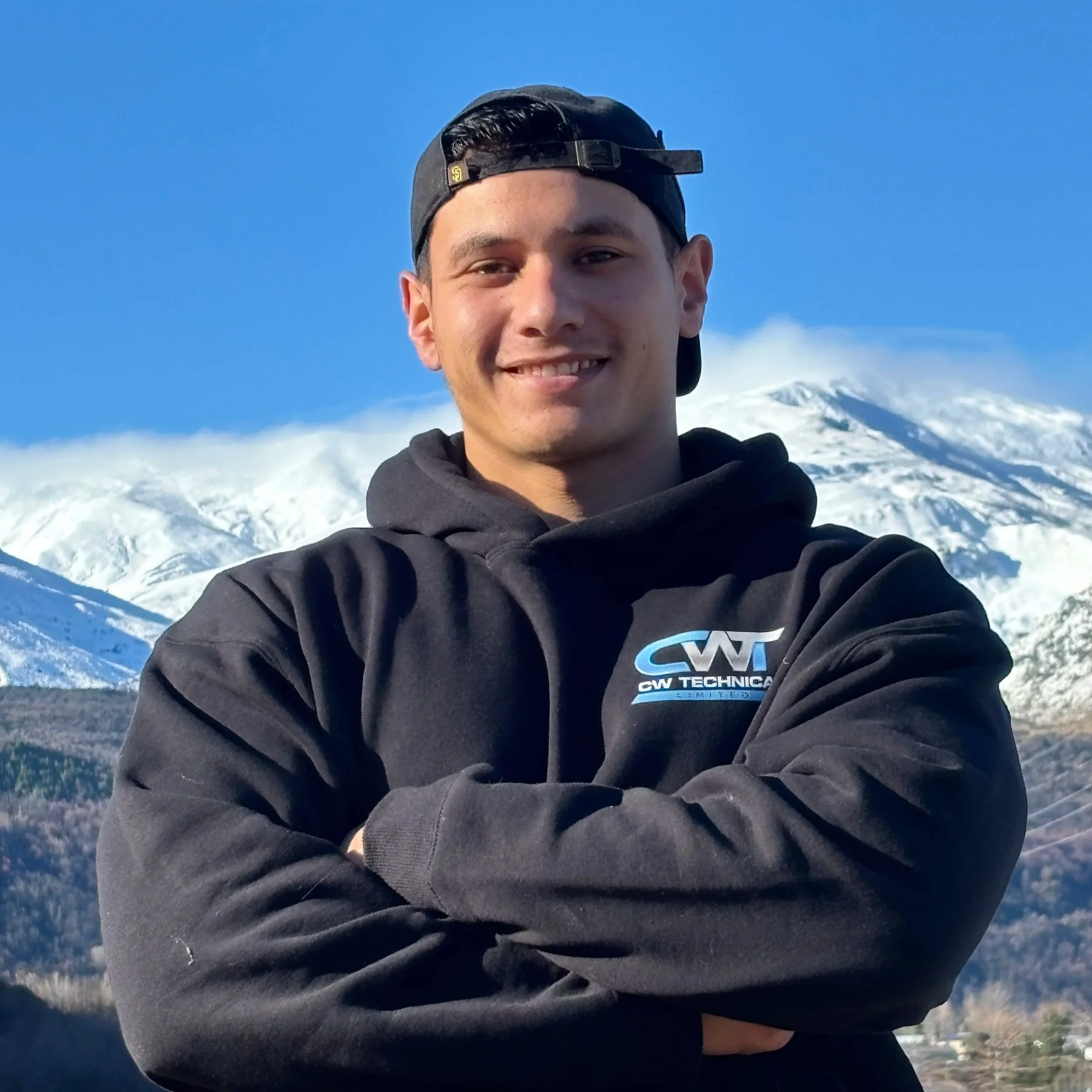 Young man smiling outdoors on a snowy mountain with clear blue sky, wearing a black cap backward and a black CW Technical hoodie.