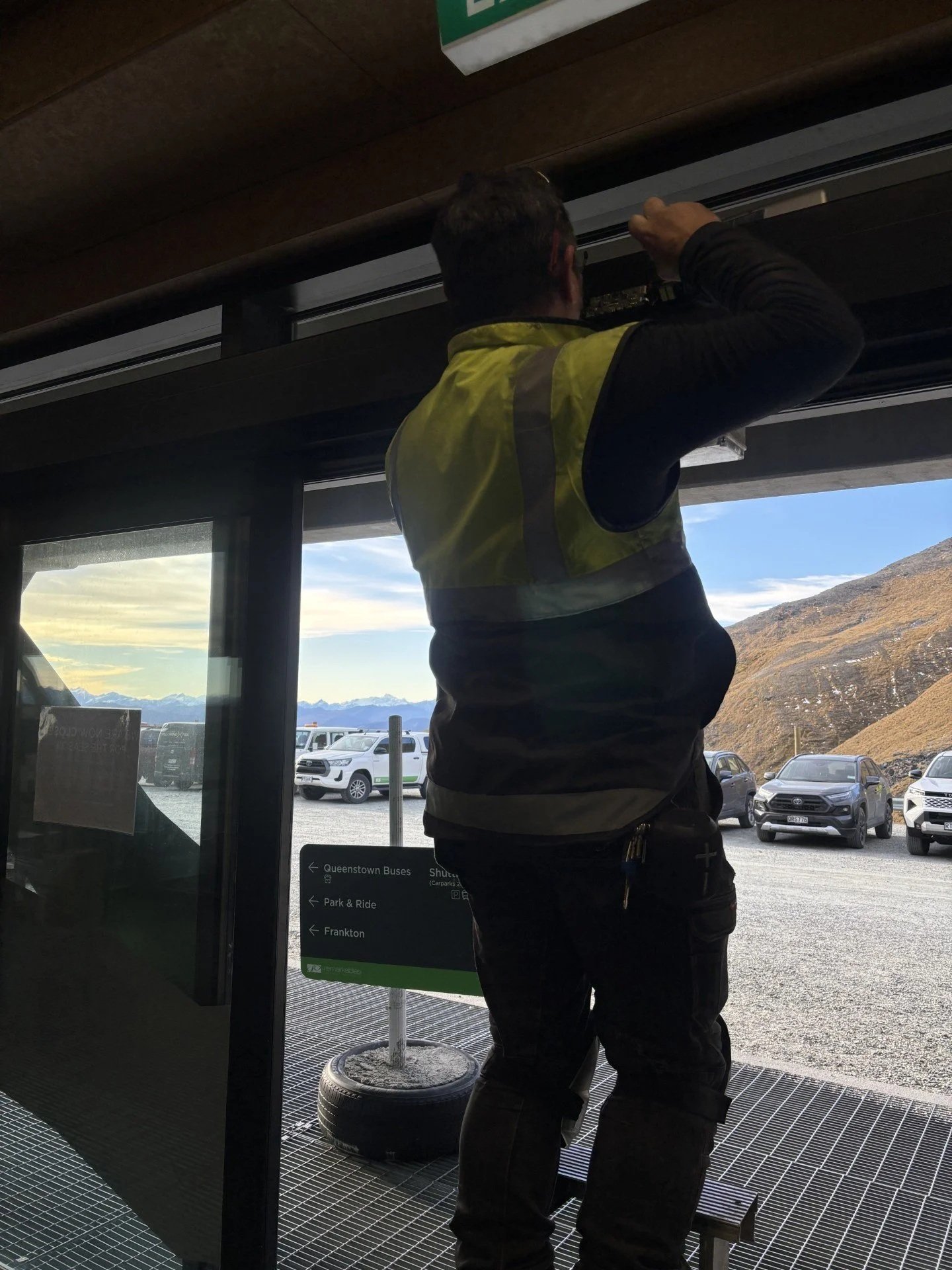 A man wearing a yellow safety vest adjusting equipment at an indoor location near a large Auto door showing a parking lot and mountain landscape.