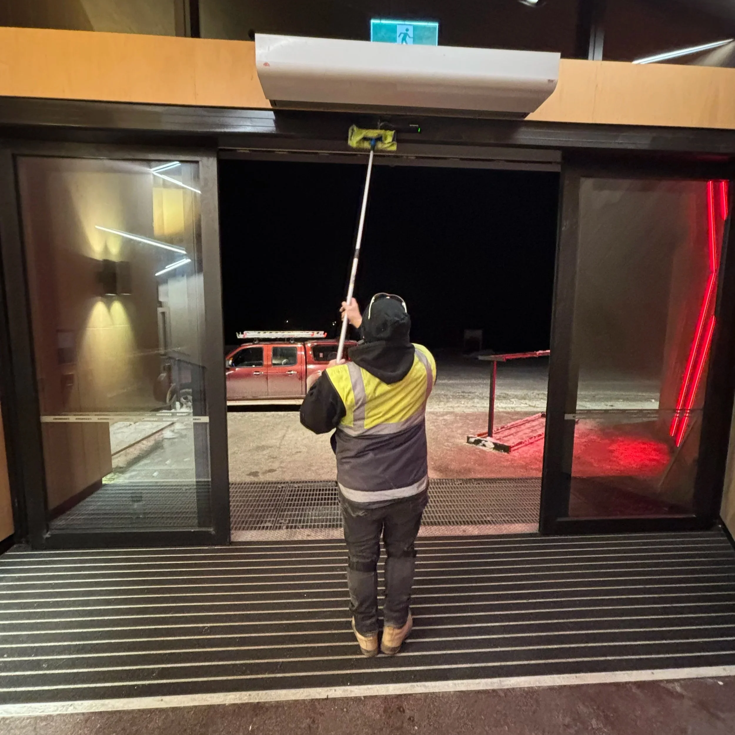 Person standing outside at night, holding a long cleaning tool, near a glass building entrance with a red vehicle parked nearby.