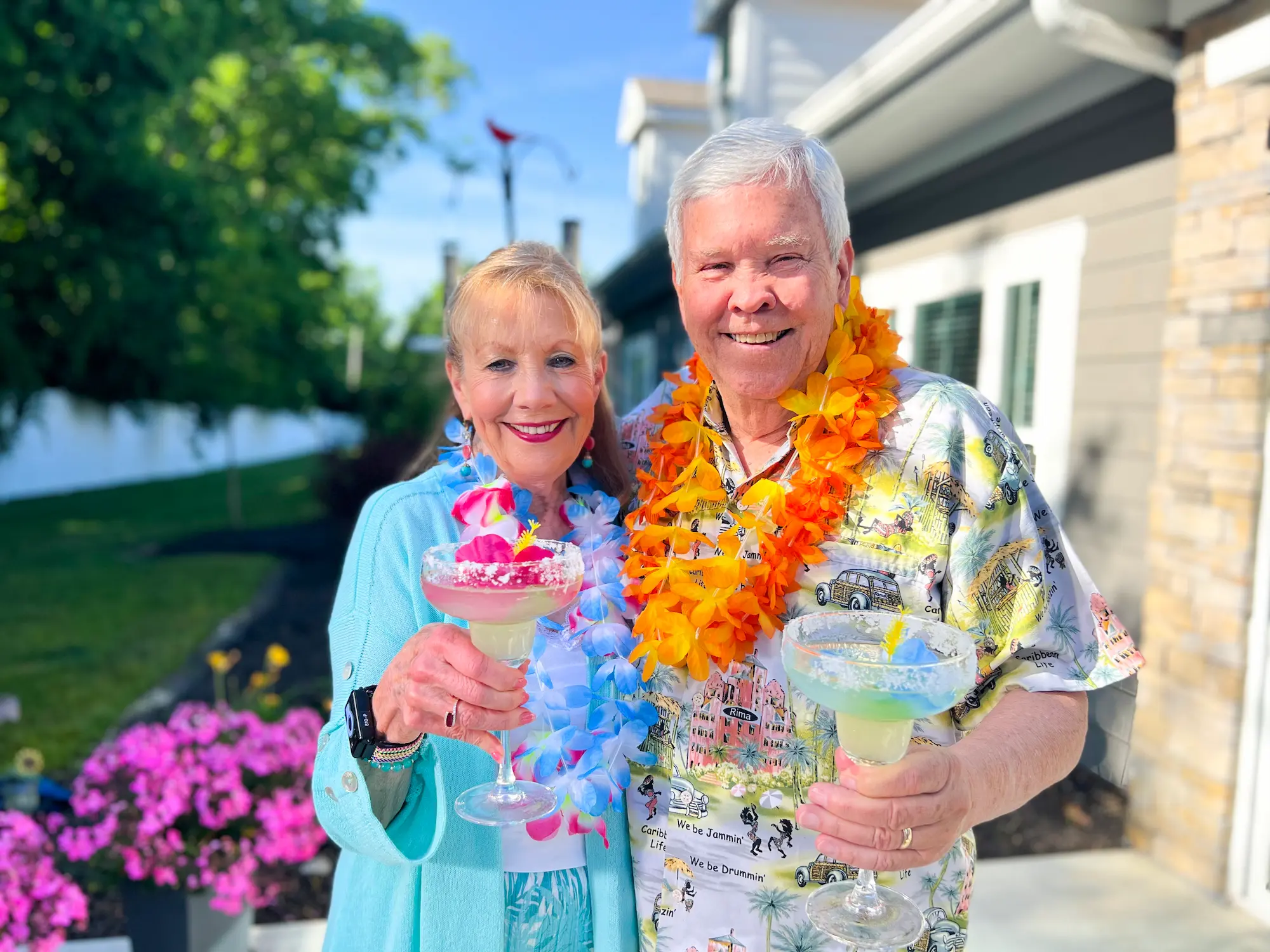 An elderly woman and man smiling outdoors, holding colorful tropical drinks with umbrellas, wearing leis, in front of a house with greenery and pink flowers.