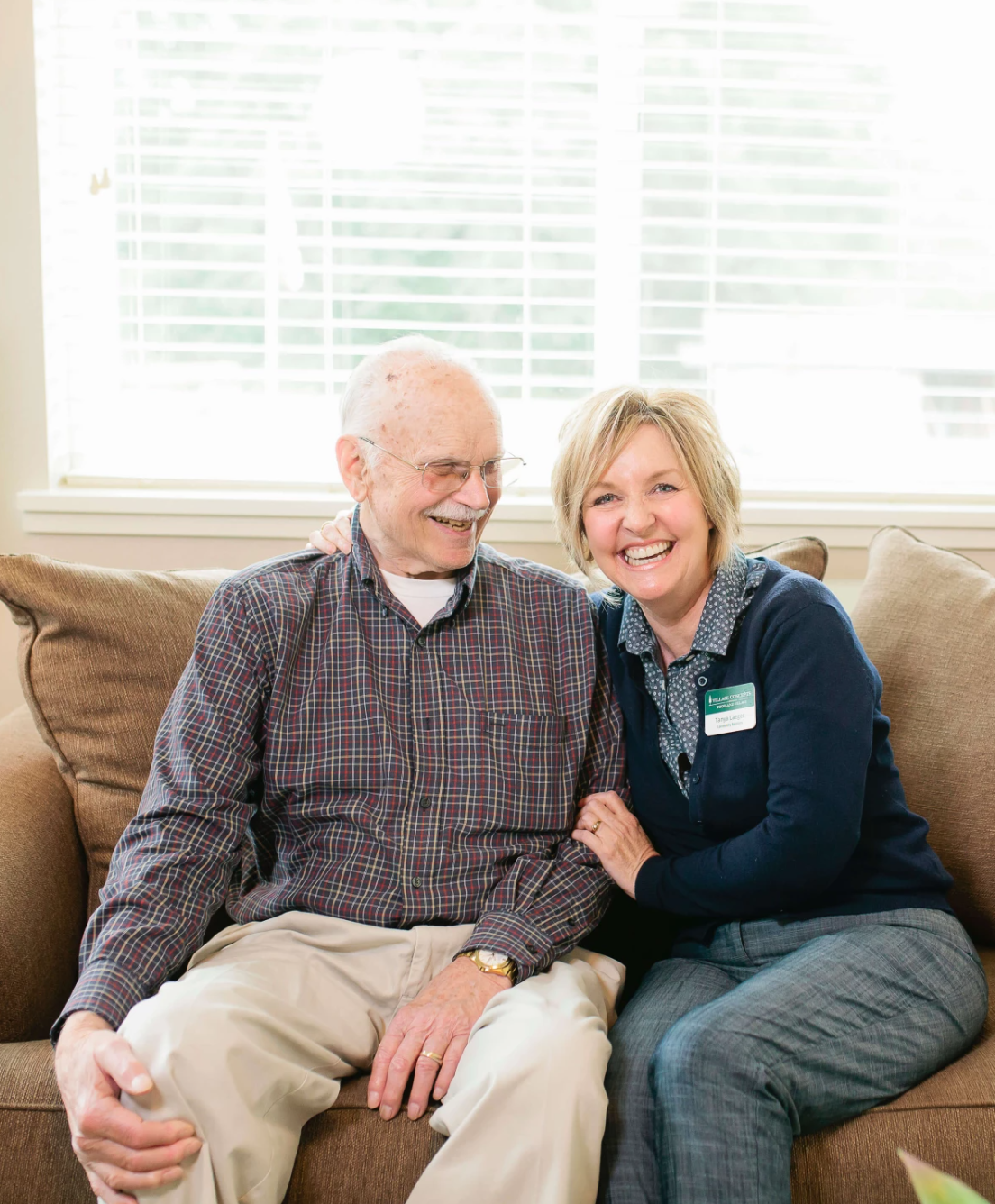 An elderly man and a woman sitting on a brown couch, smiling and enjoying each other's company, with a bright window in the background.