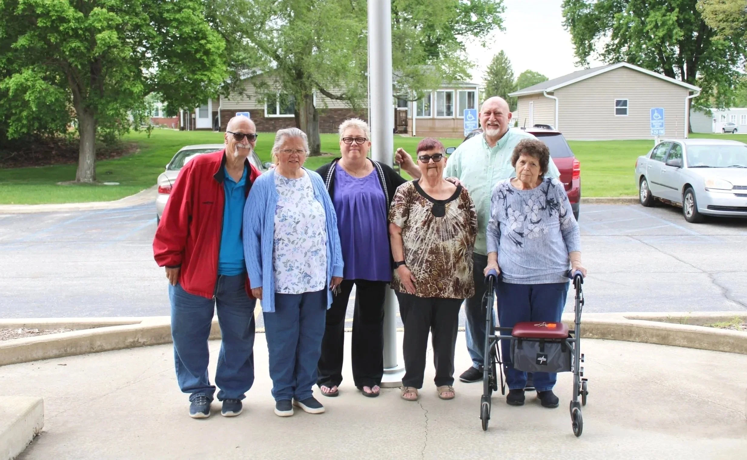 Group of seven senior adults standing outdoors in a parking lot, with some smiling and one using a walker, in front of trees and residential houses.