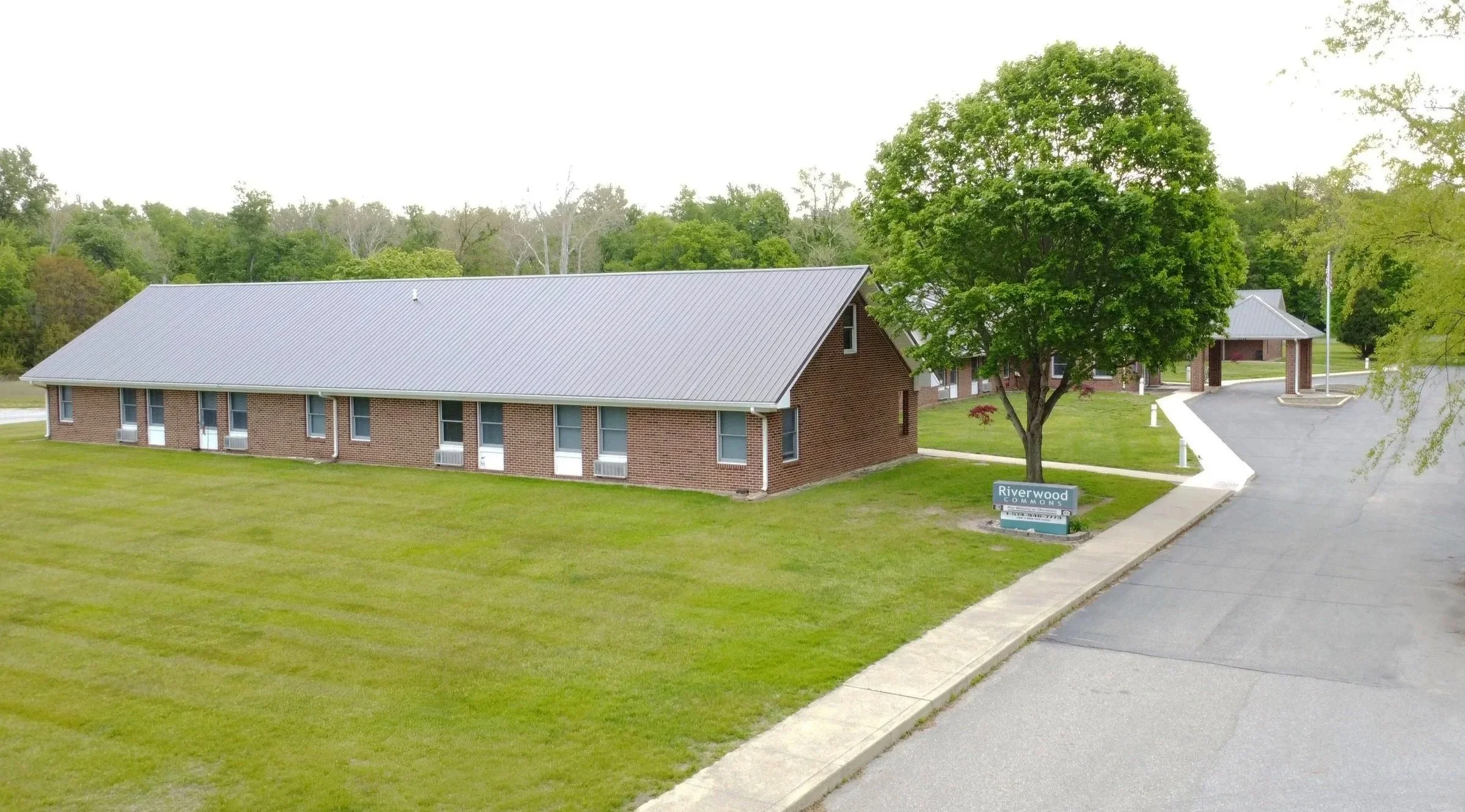 A brick building with a metal roof surrounded by a well-maintained lawn and trees, with a sign that reads 'Riverwood Commons' near a curved paved road.