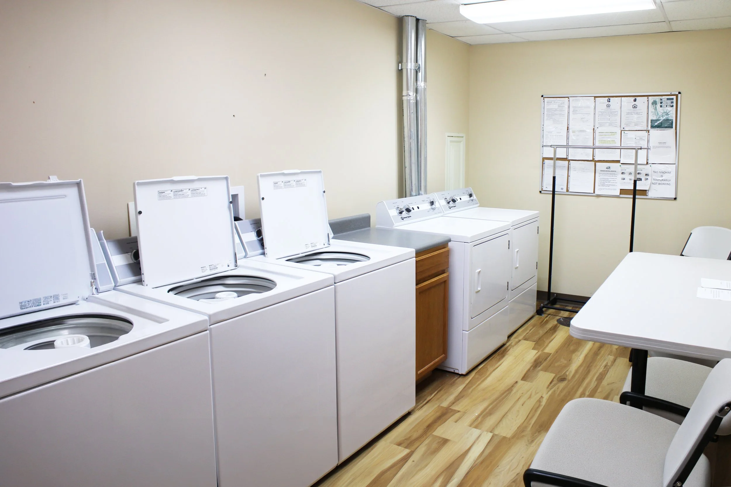 laundry room with four washing machines and one dryer, bulletin board on the wall, white table, white walls, wood-patterned vinyl flooring.