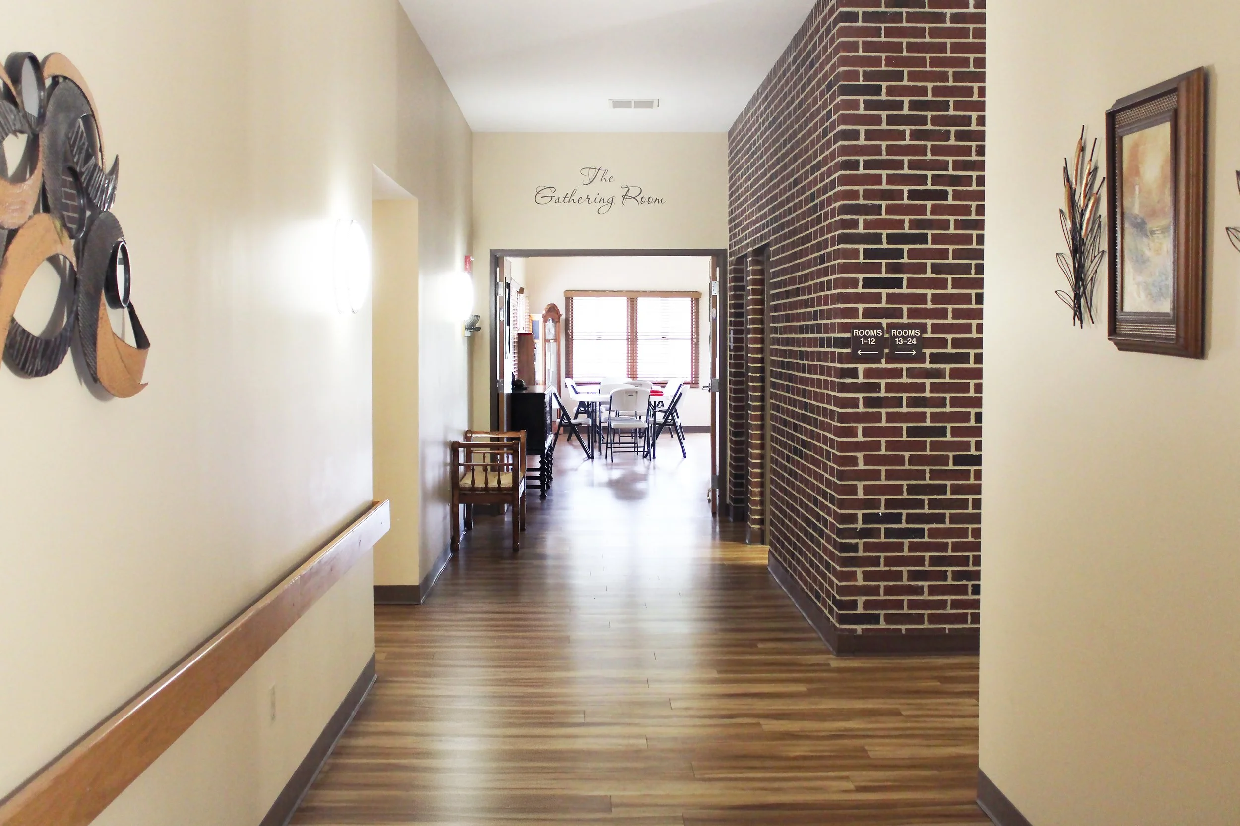 Hallway leading to a dining area with a table and chairs, brick wall on the right, and a sign indicating room numbers 1-12 and 13-24.