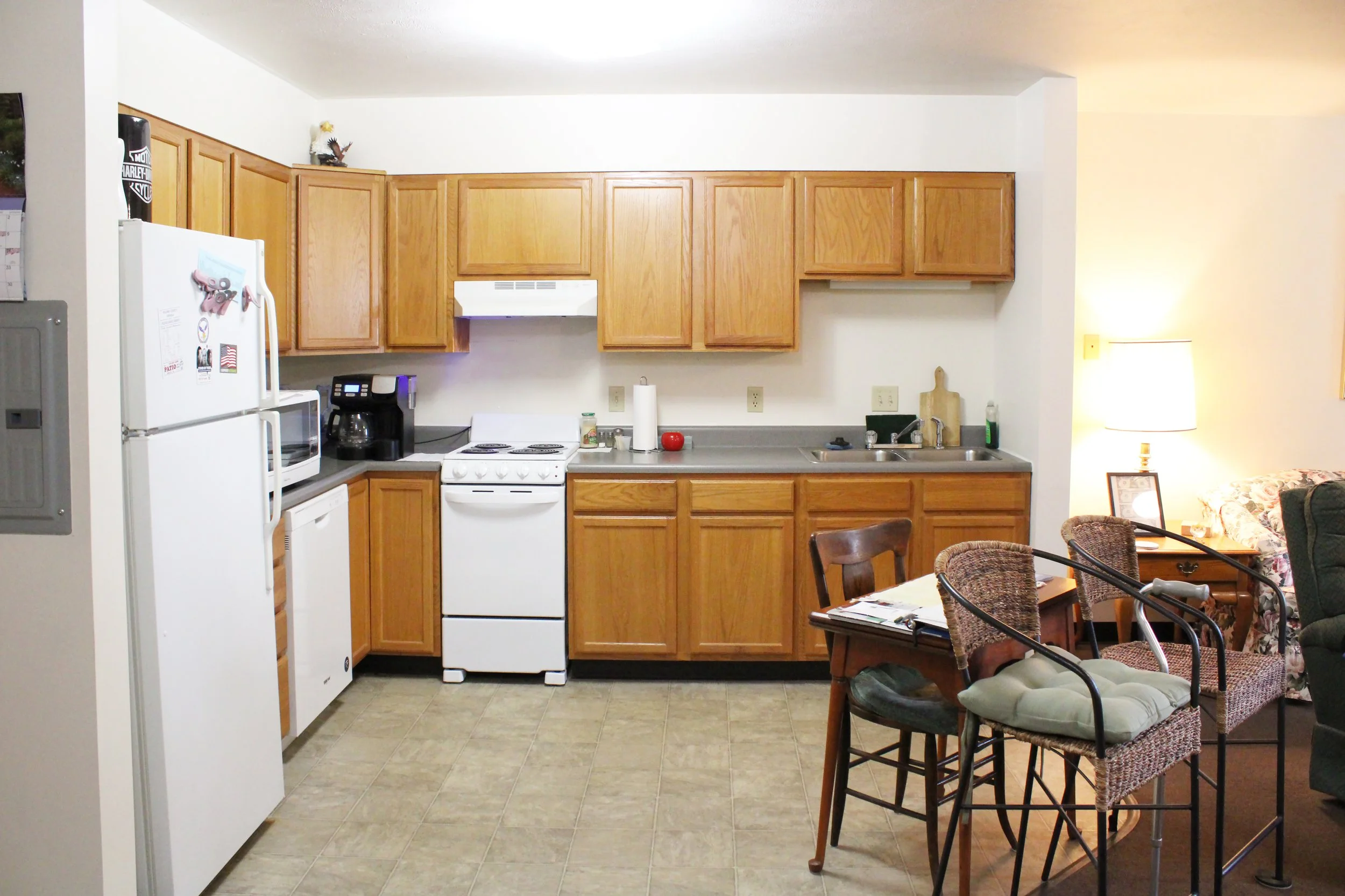 A kitchen and dining area with wooden cabinets, white appliances, and a counter with a paper towel holder. There's a small table with chairs and a living room area with a lamp and sofa visible.