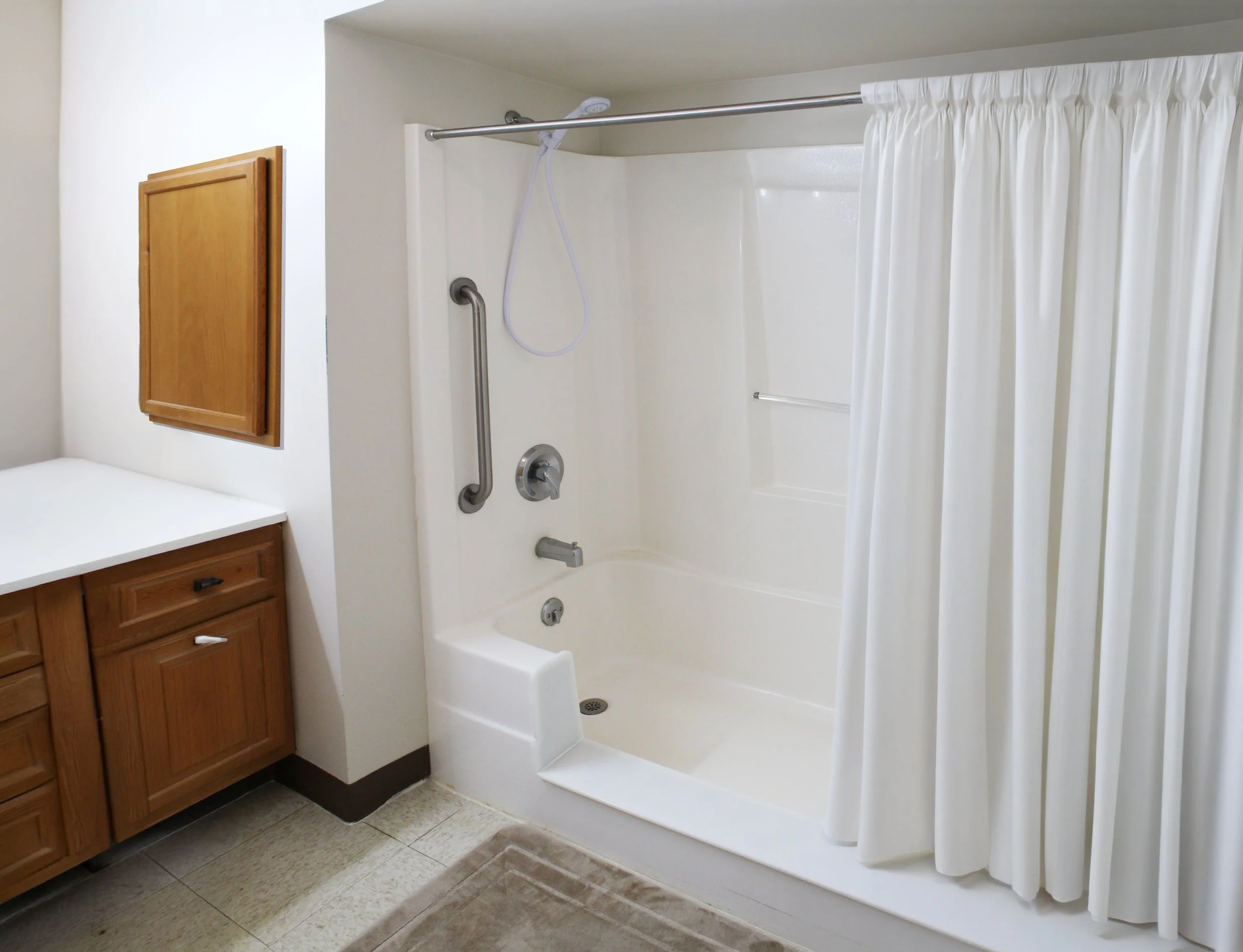 A bathroom shower with a white curtain, grab bars, a hand shower, and shower controls. There are wooden cabinets to the left and a tiled floor.