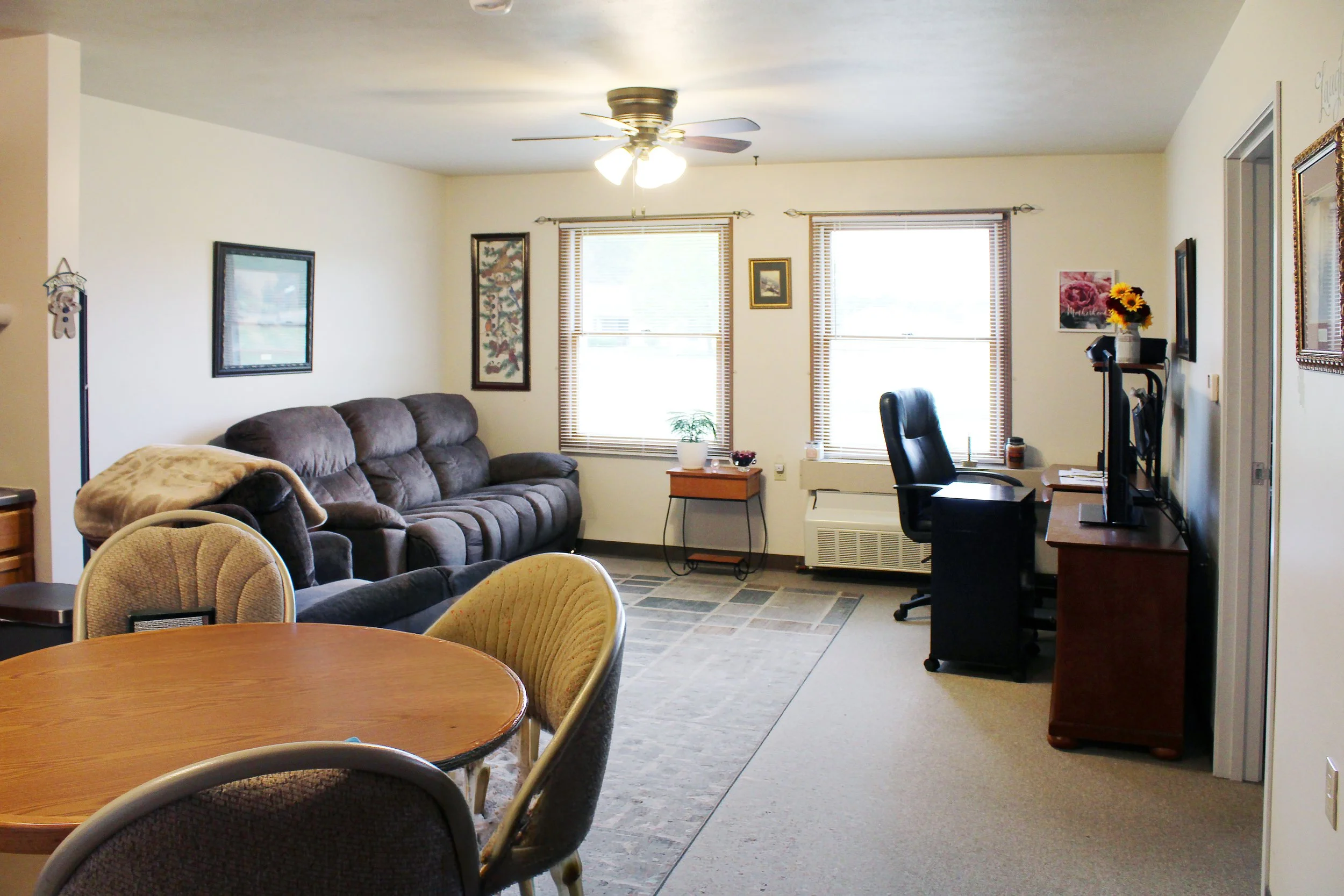 A living room with a brown couch, a small side table, and windows with wooden blinds. There are framed pictures on the walls, a ceiling fan with lights, and a desk with a computer monitor and a black swivel chair. The room has a mix of natural and ar
