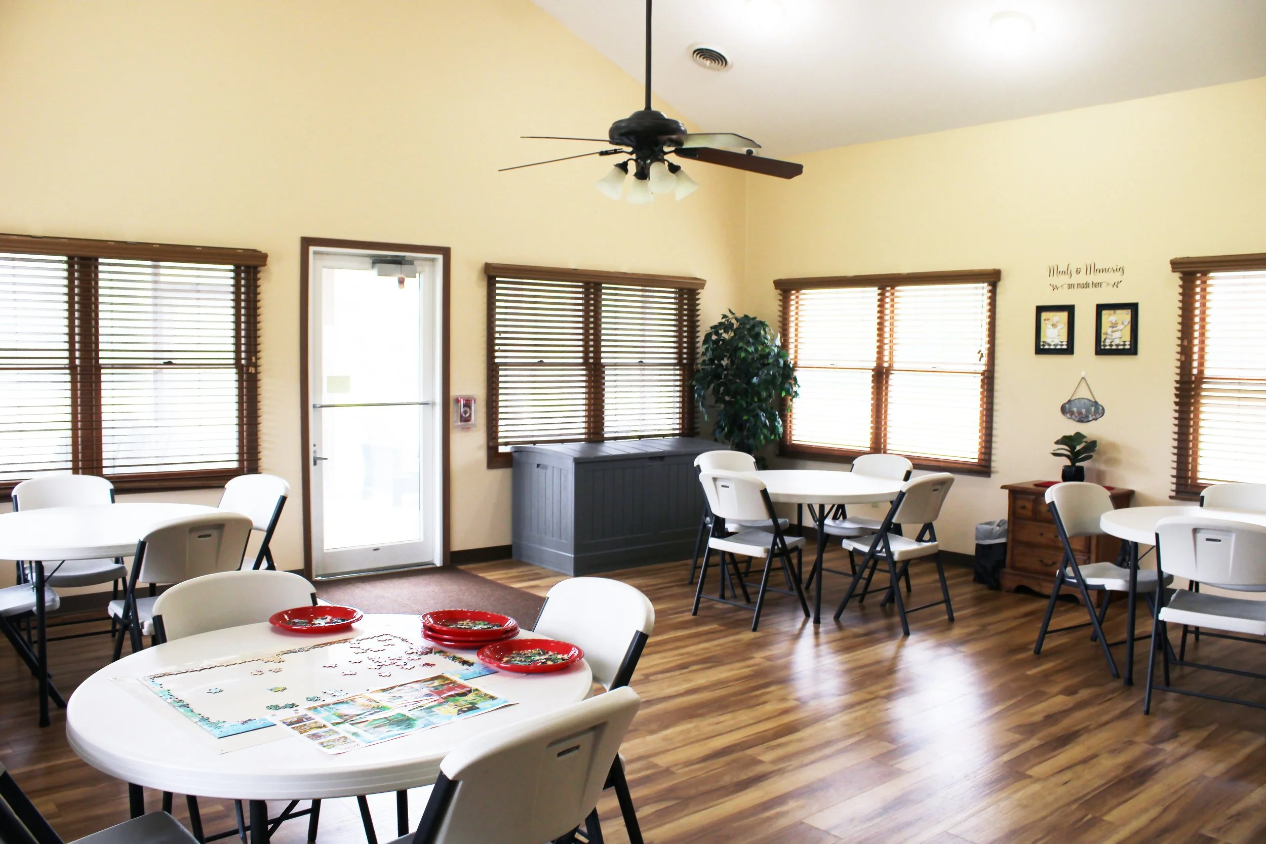 A bright room with wooden floors, multiple windows with wooden blinds, and a door. There are several white chairs and round tables. A ceiling fan with light fixtures hangs from the ceiling. Decor includes framed pictures, a potted plant, and a small 