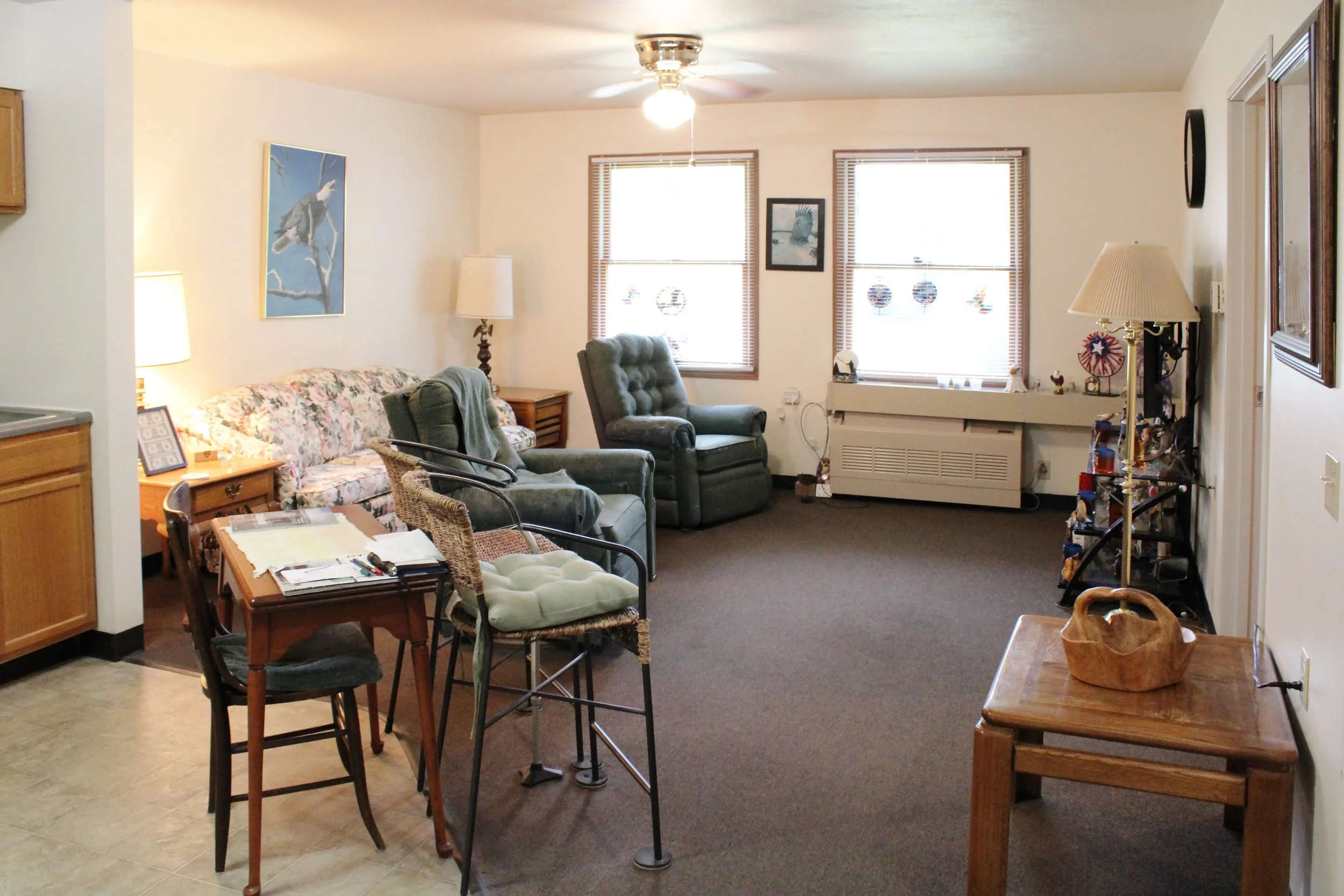 Living room with three large windows, a ceiling fan with light, a floral sofa, two green armchairs, a wooden side table, and various framed pictures and decorations on the walls.