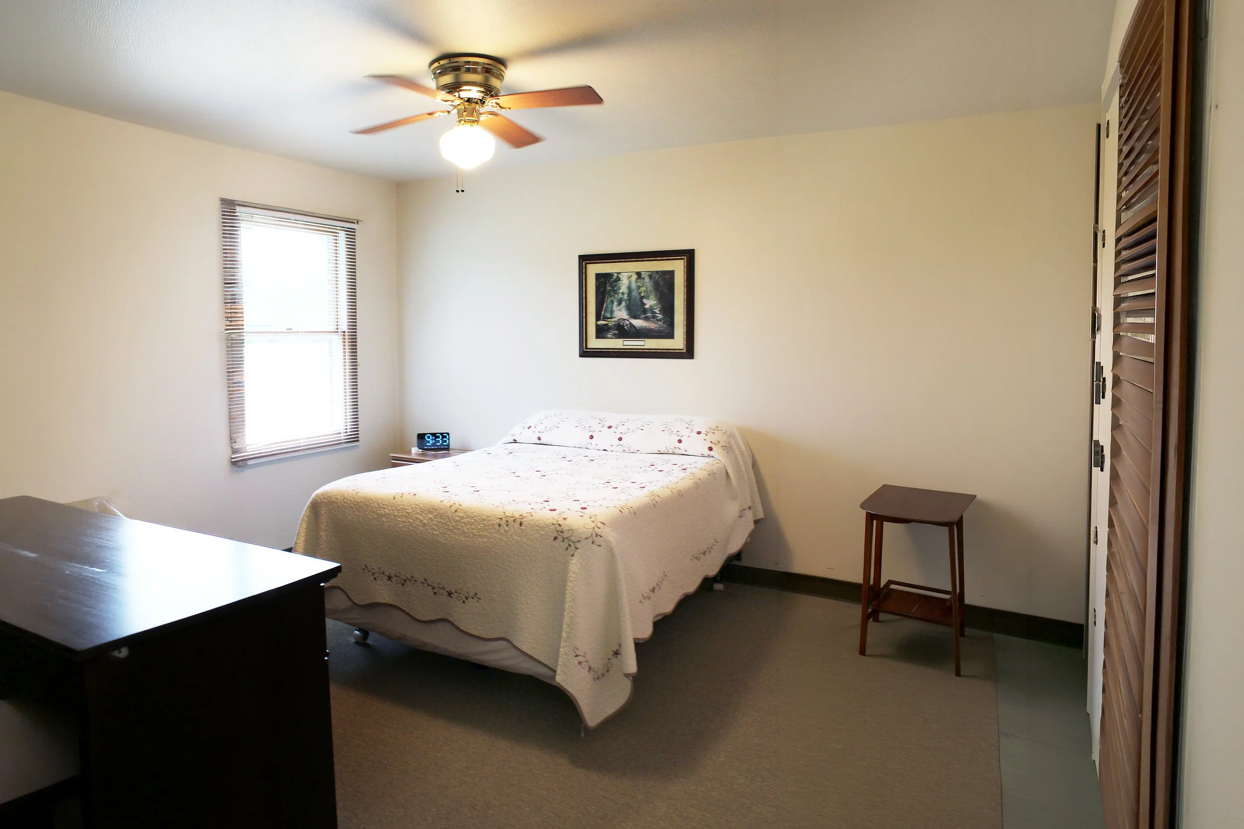 A cozy bedroom with a bed covered in a white quilt with pink floral embroidery. There is a small wooden side table next to the bed. Two windows with blinds are on the wall, allowing natural light into the room. A framed picture of a forest scene hang