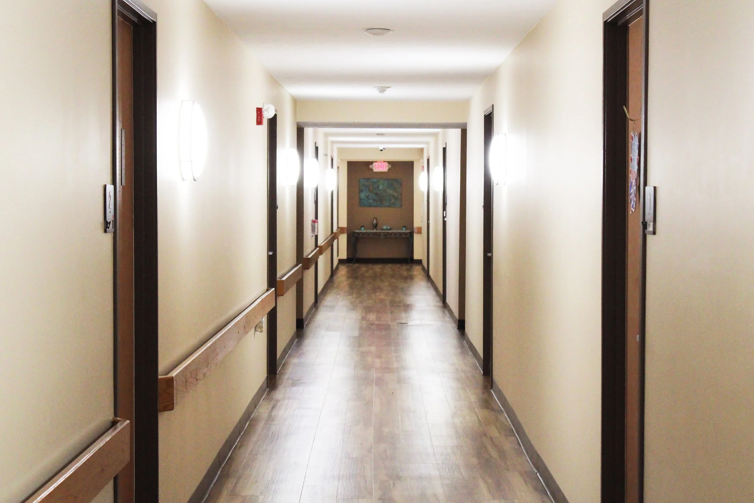 Empty hotel corridor with closed doors on either side, light fixtures on the walls, and a table with a decorative item at the end of the hallway.