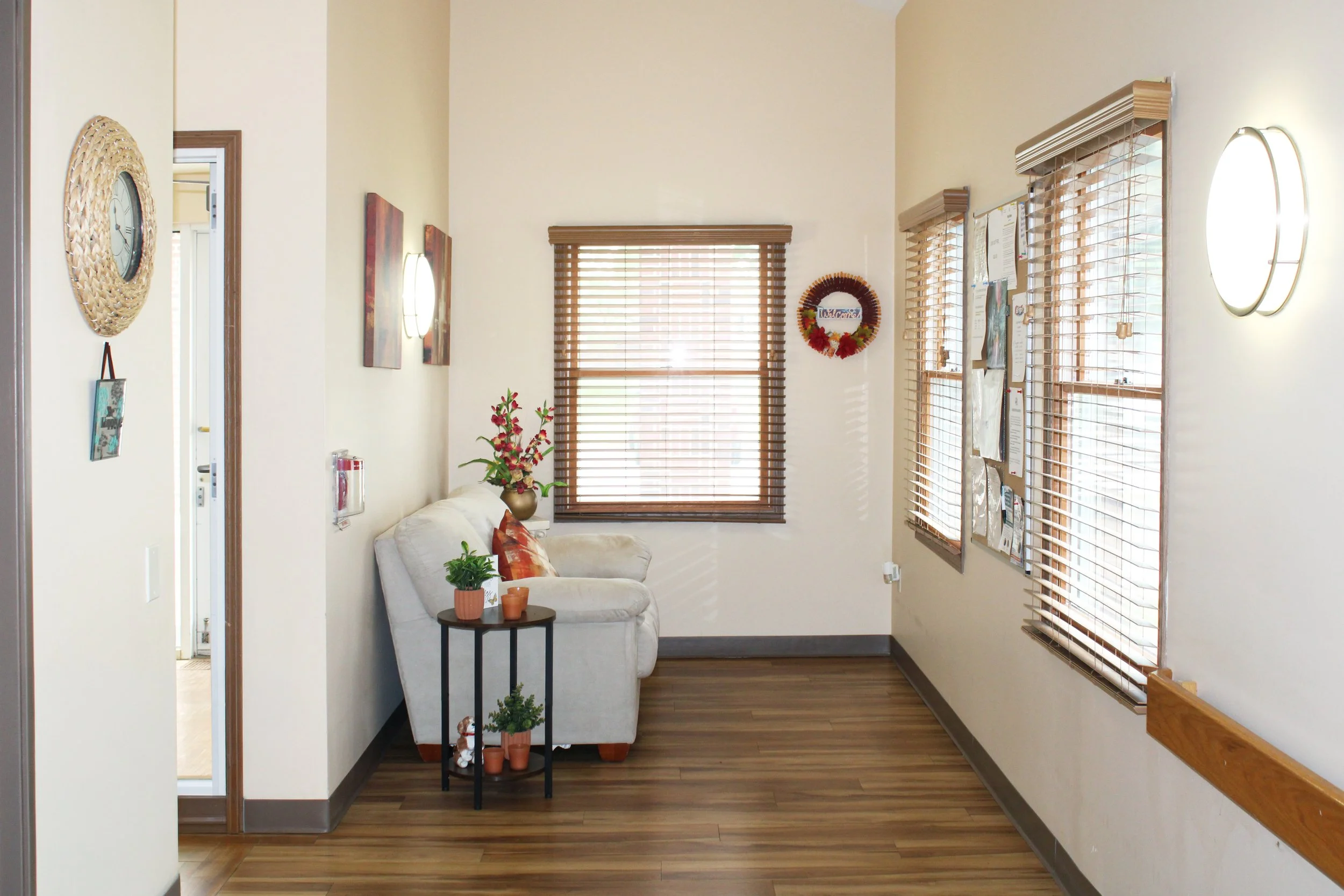 Interior view of a cozy living room with beige walls, wooden blinds on two large windows, a small sofa with decorative pillows, a side table with plants and a small stuffed animal, wall-mounted lights, and decorative wall hangings.