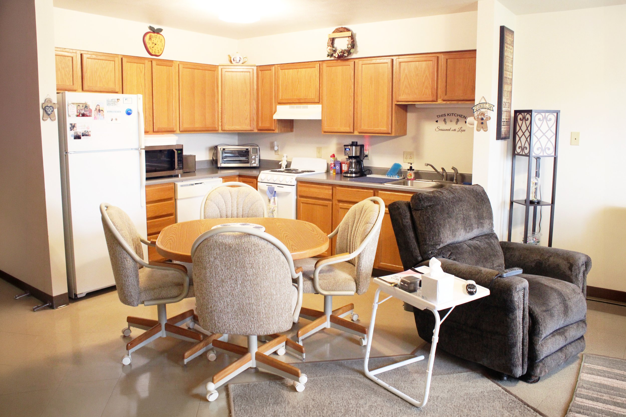 A kitchen with wooden cabinets, a white refrigerator with magnets, a stove, and small appliances, adjacent to a dining area with a round table and chairs. There is a brown sofa and a side table with remote controls and a tissue box in the living area