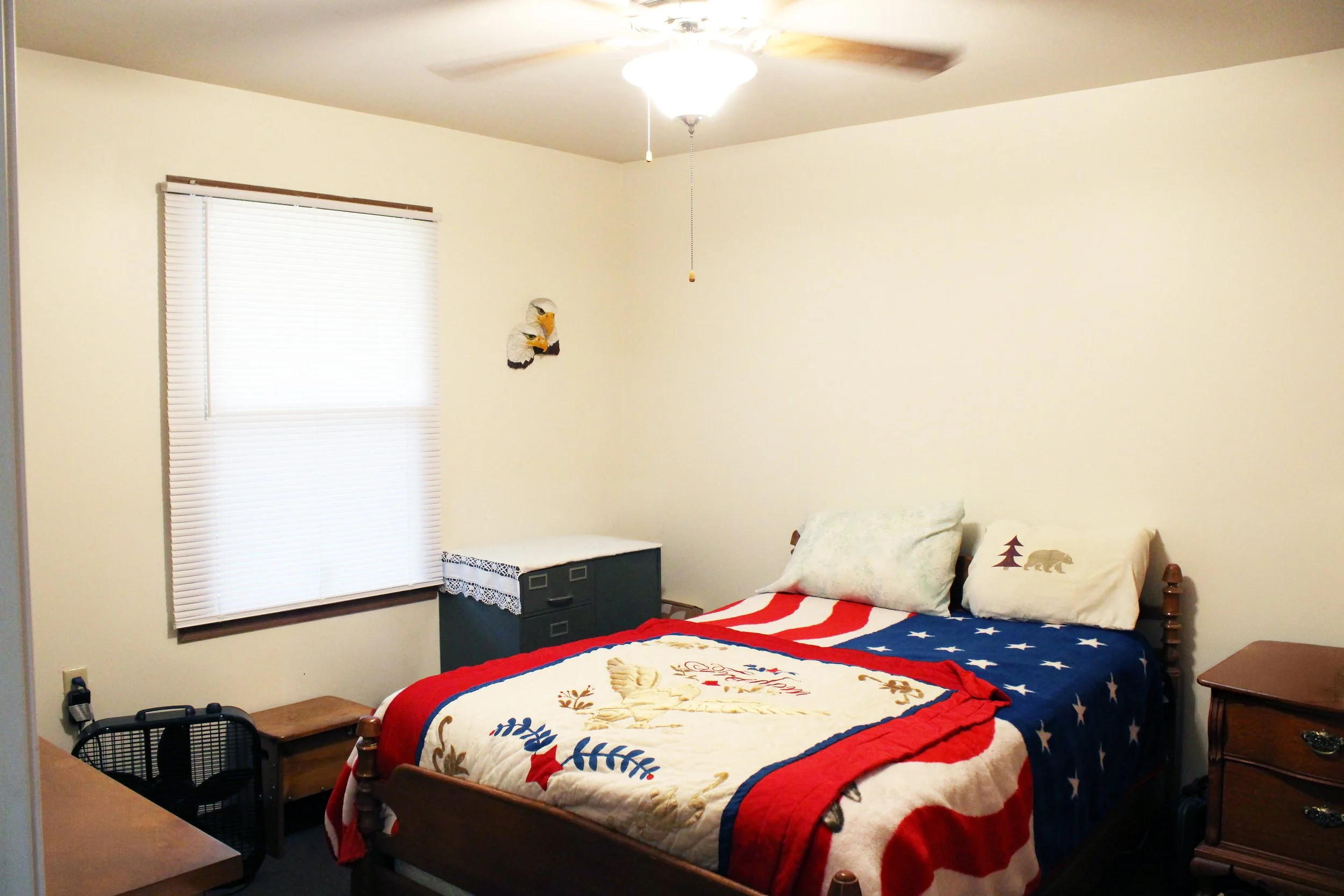 A bedroom with a bed covered in a patriotic-themed quilt and pillows featuring a bear and trees. The room has a window with closed blinds, a ceiling fan with light, and a small dresser or nightstand.