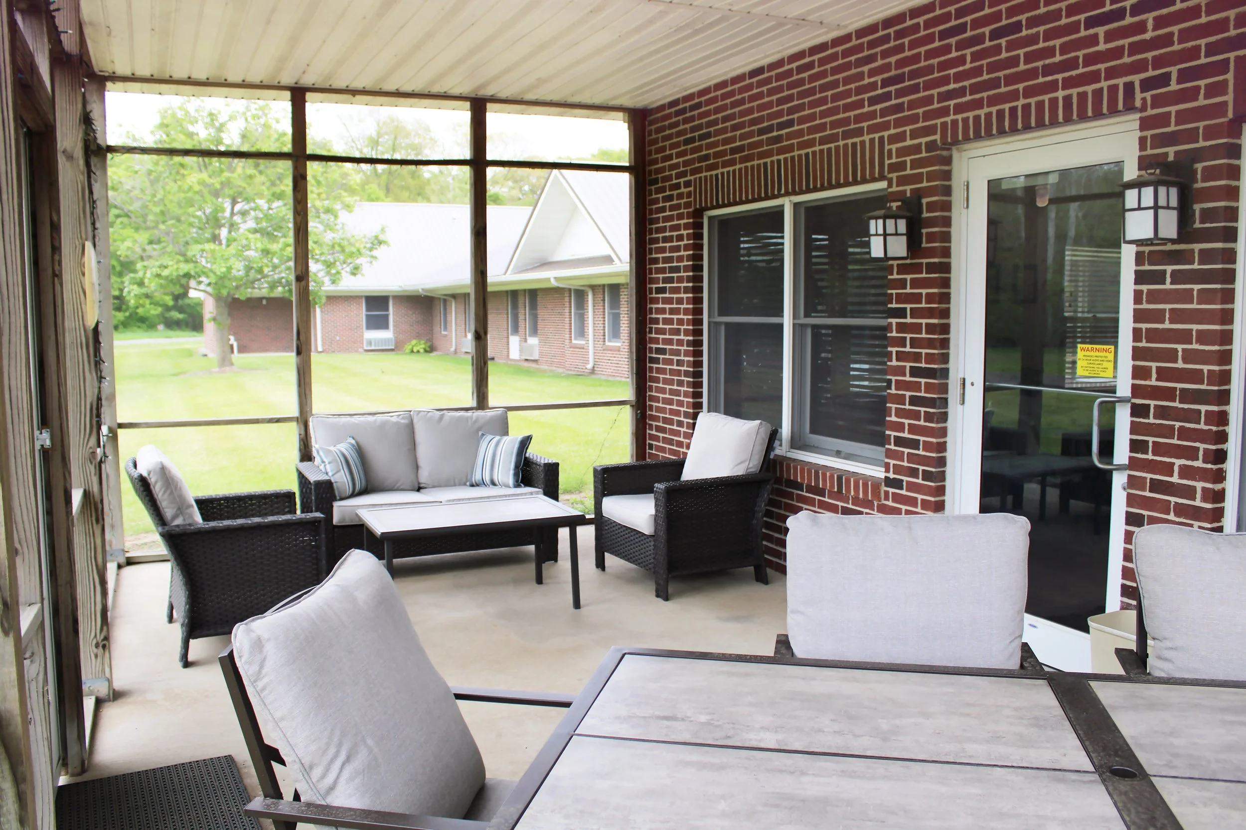 Covered outdoor patio with black wicker furniture, including sofas and chairs with cushions, next to a brick house with sliding glass door and wall-mounted lanterns, overlooking a lawn and neighboring houses.