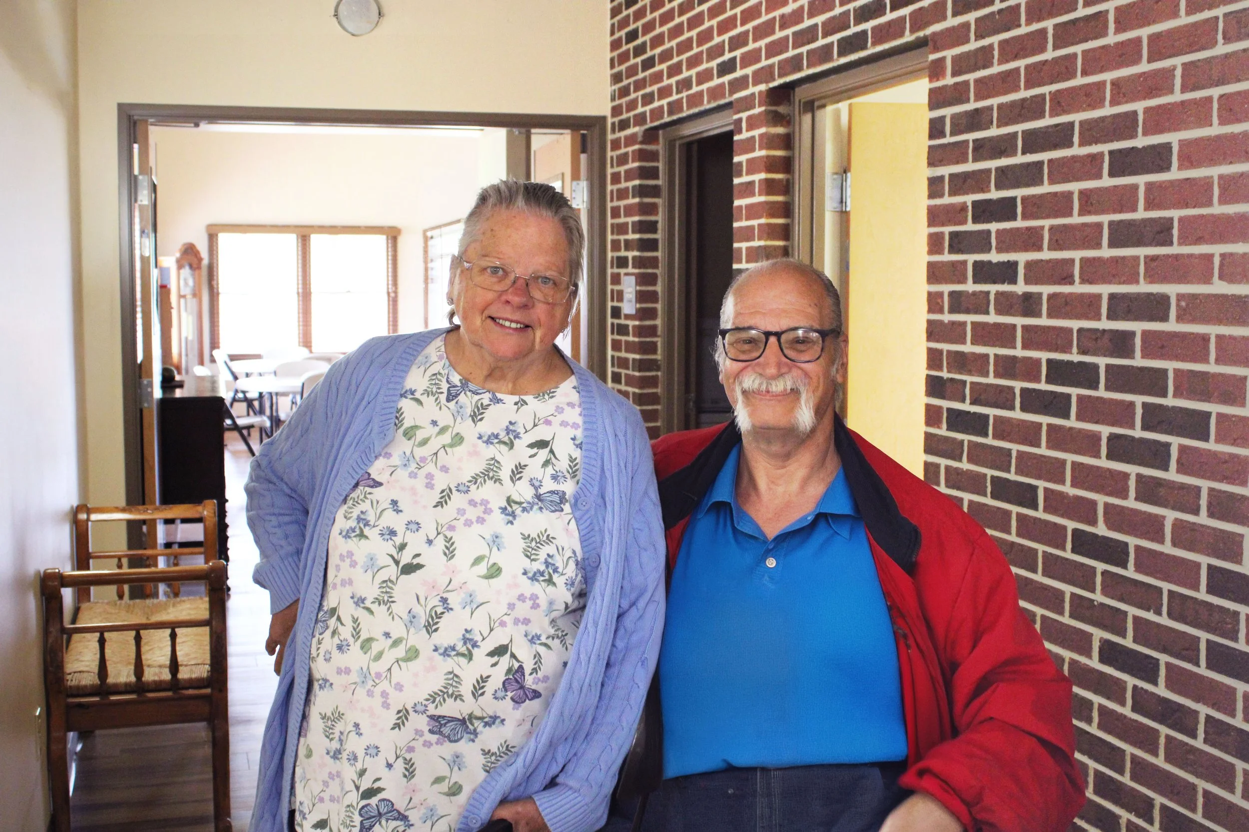 An elderly woman with glasses and a floral blouse smiling next to an elderly man with glasses and a red jacket in a hallway with a brick wall.