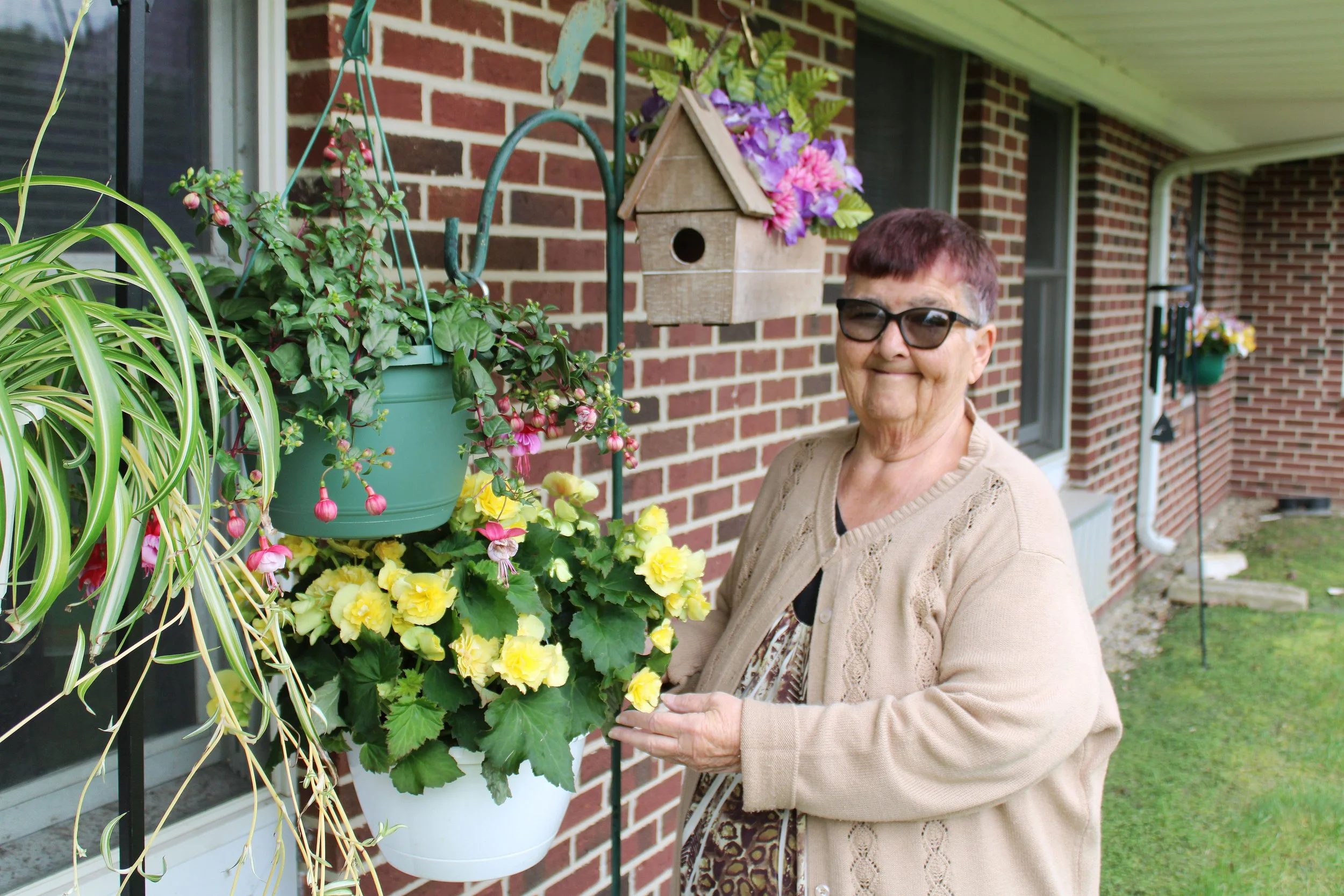 An elderly woman with short purple hair and glasses standing on a porch near hanging flower baskets, a birdhouse, and a brick house wall.