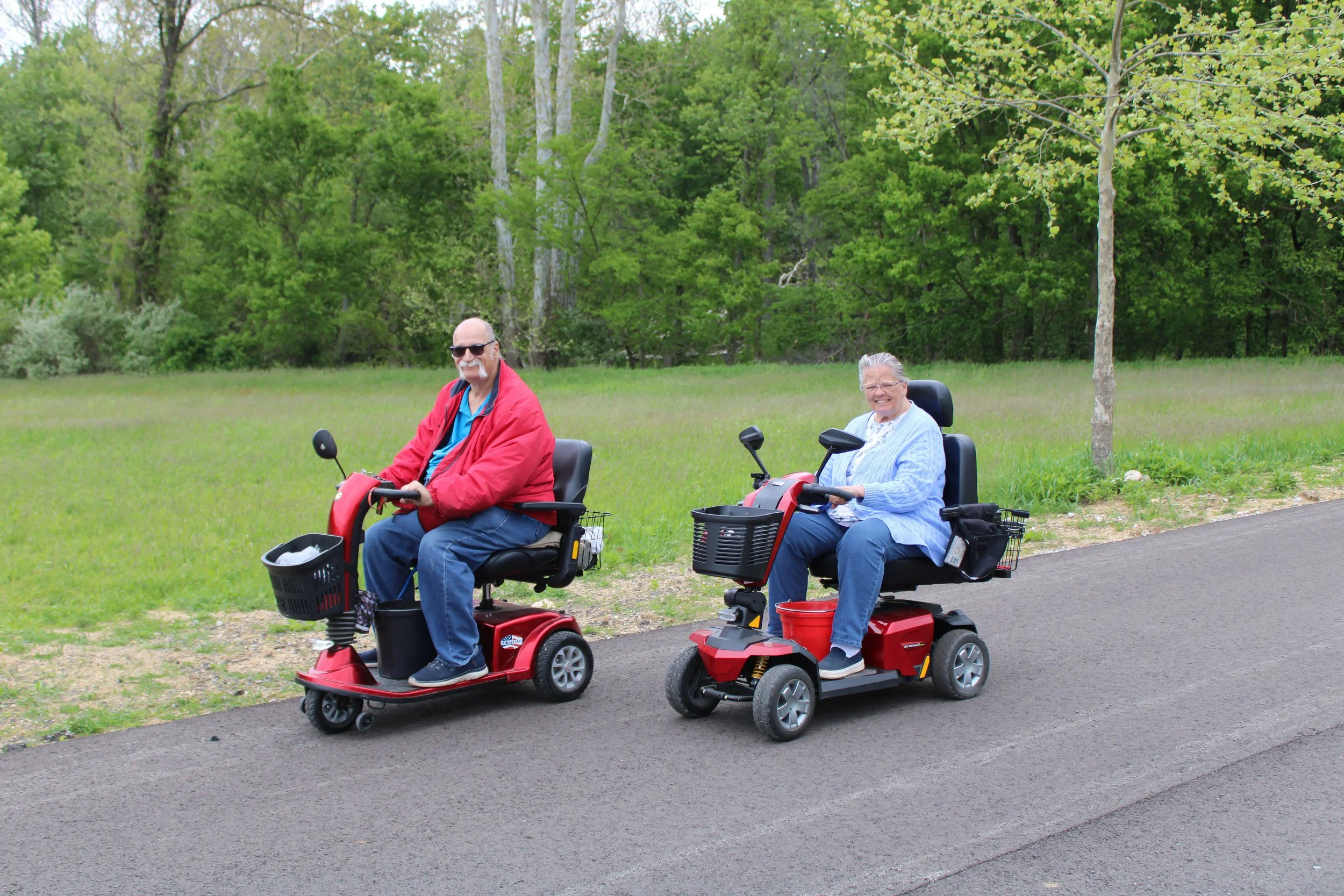 Two elderly individuals, a man and a woman, riding mobility scooters on a paved path in a park with green grass and trees.