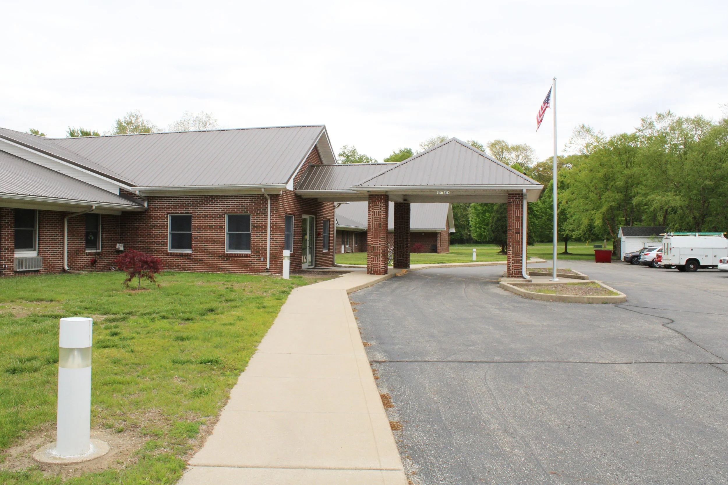 A brick building with a gray metal roof, a covered entrance, and a flagpole with an American flag. There is a sidewalk and parking lot with a few cars and a utility truck. Green trees are in the background under a cloudy sky.