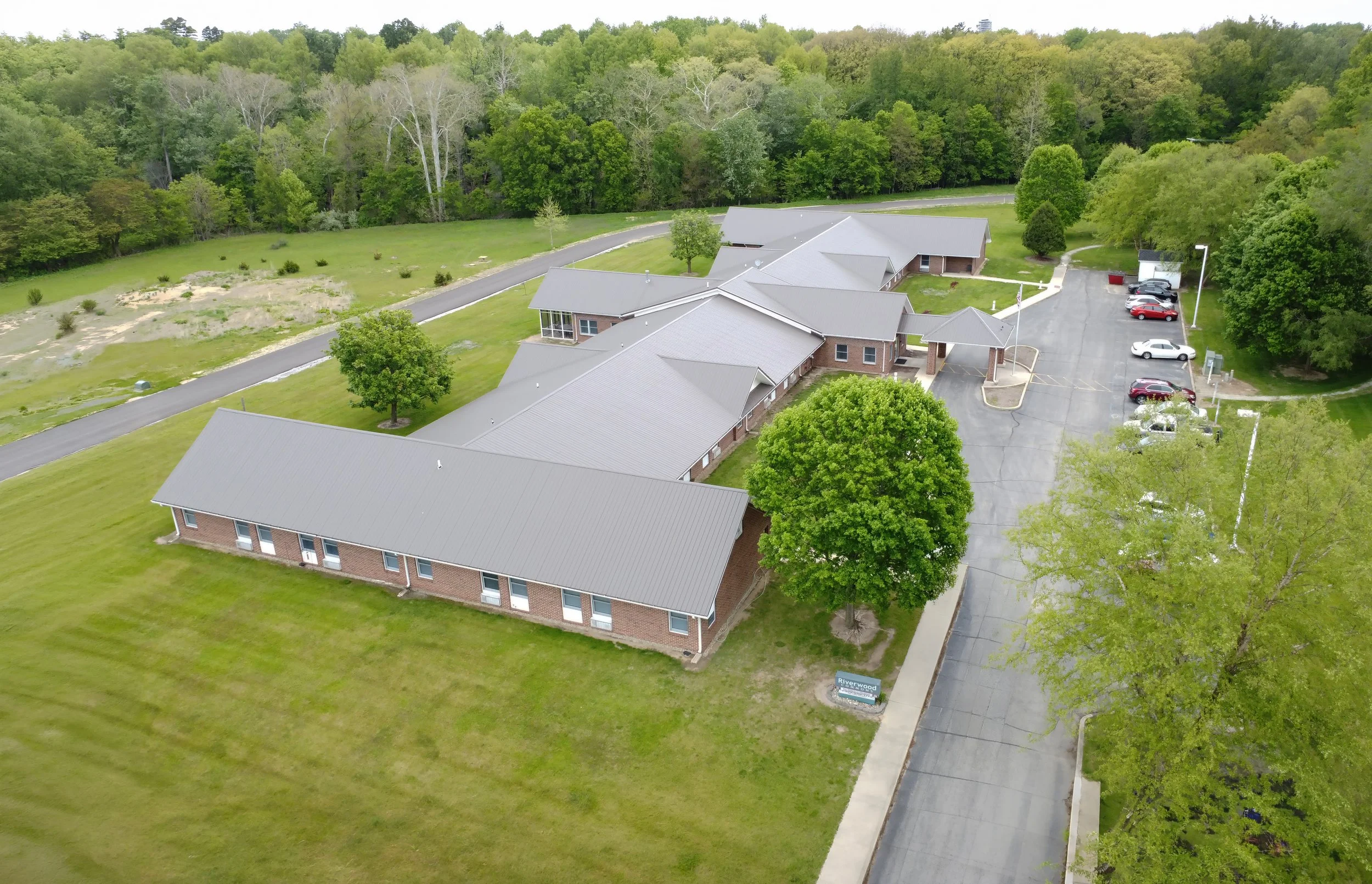 Aerial view of a building complex surrounded by green trees and grassy areas, with a parking lot to the right side and a driveway leading to the entrance.