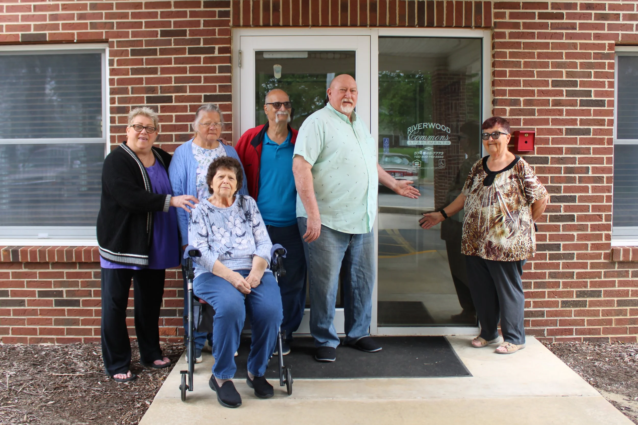 Group of six elderly people, some with disabilities, standing in front of an apartment building entrance. One woman is in a wheelchair, others are standing and smiling.