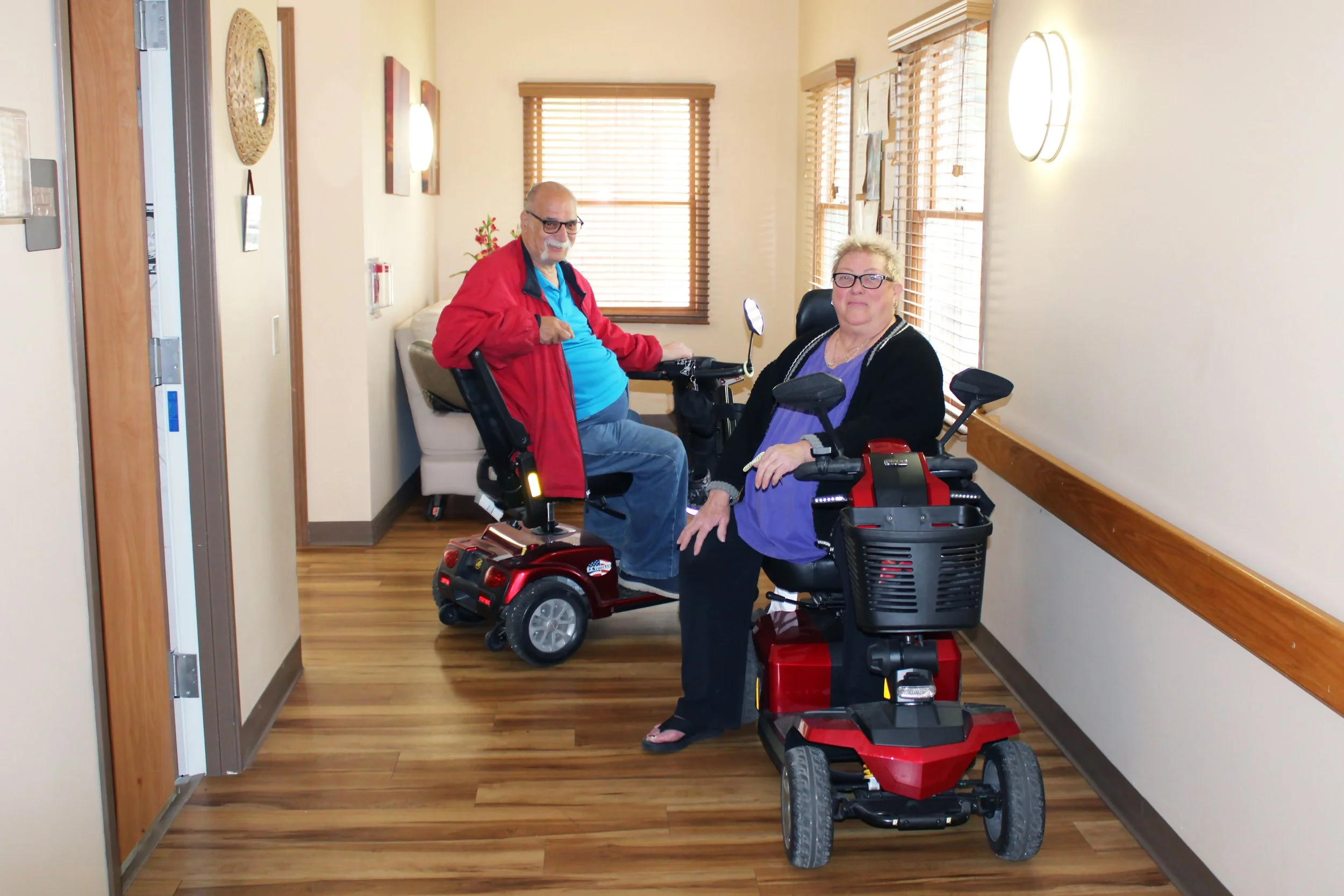 An elderly man and woman with mobility aids in a hospital corridor, smiling at the camera.