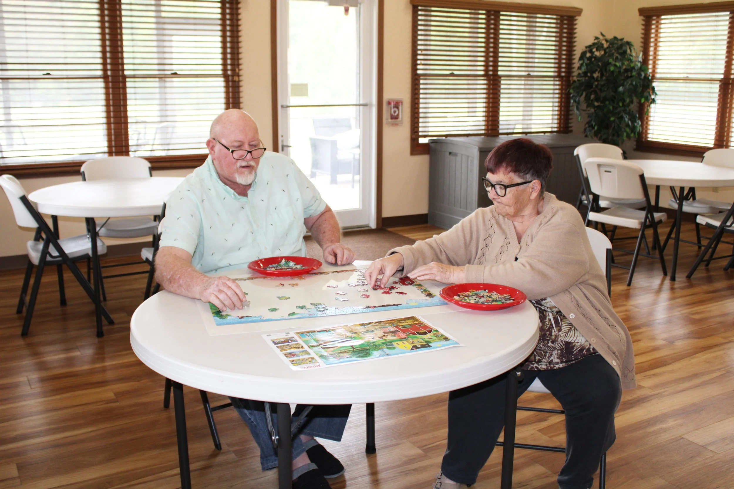 Two elderly women sitting at a round table working on a jigsaw puzzle in a bright room with large windows, blinds, and a wooden floor.