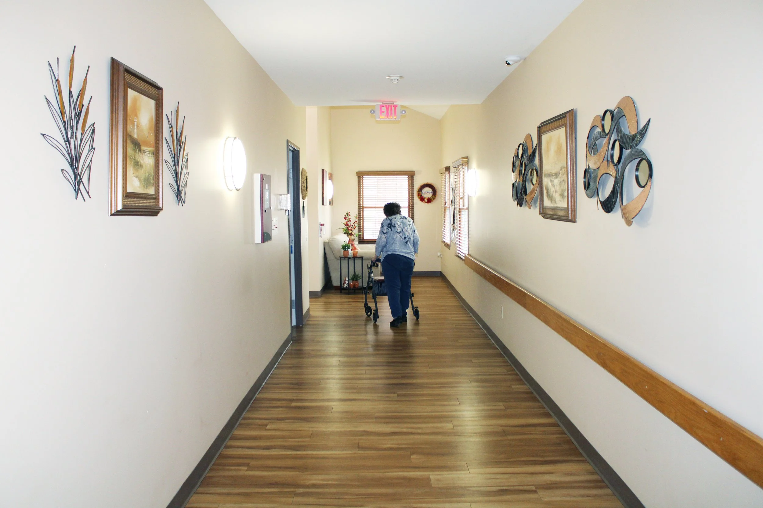A hallway with light-colored walls decorated with framed pictures and circular metal wall art. Wooden blinds cover the windows, and a person with a walker is walking away from the camera. The floor is wood, and there is an illuminated exit sign above the hallway.