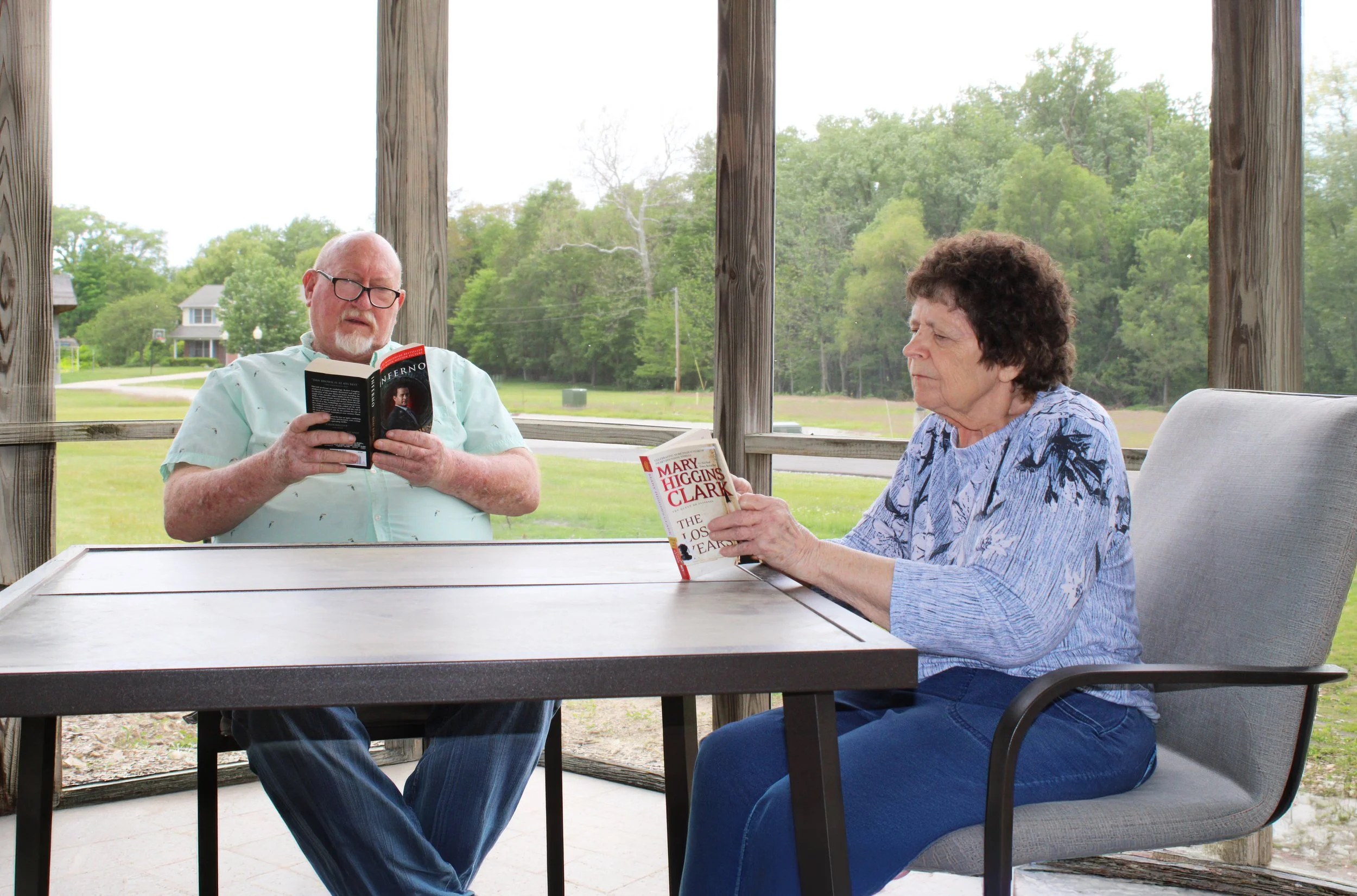 An elderly man and woman sitting at a table on a covered porch, reading books. The man is reading a book titled "Inferno" by Dan Brown, and the woman is reading a book titled "The Lost Queen" by Mary Higgins Clark. The porch has wooden posts and the background shows trees and a grassy area.