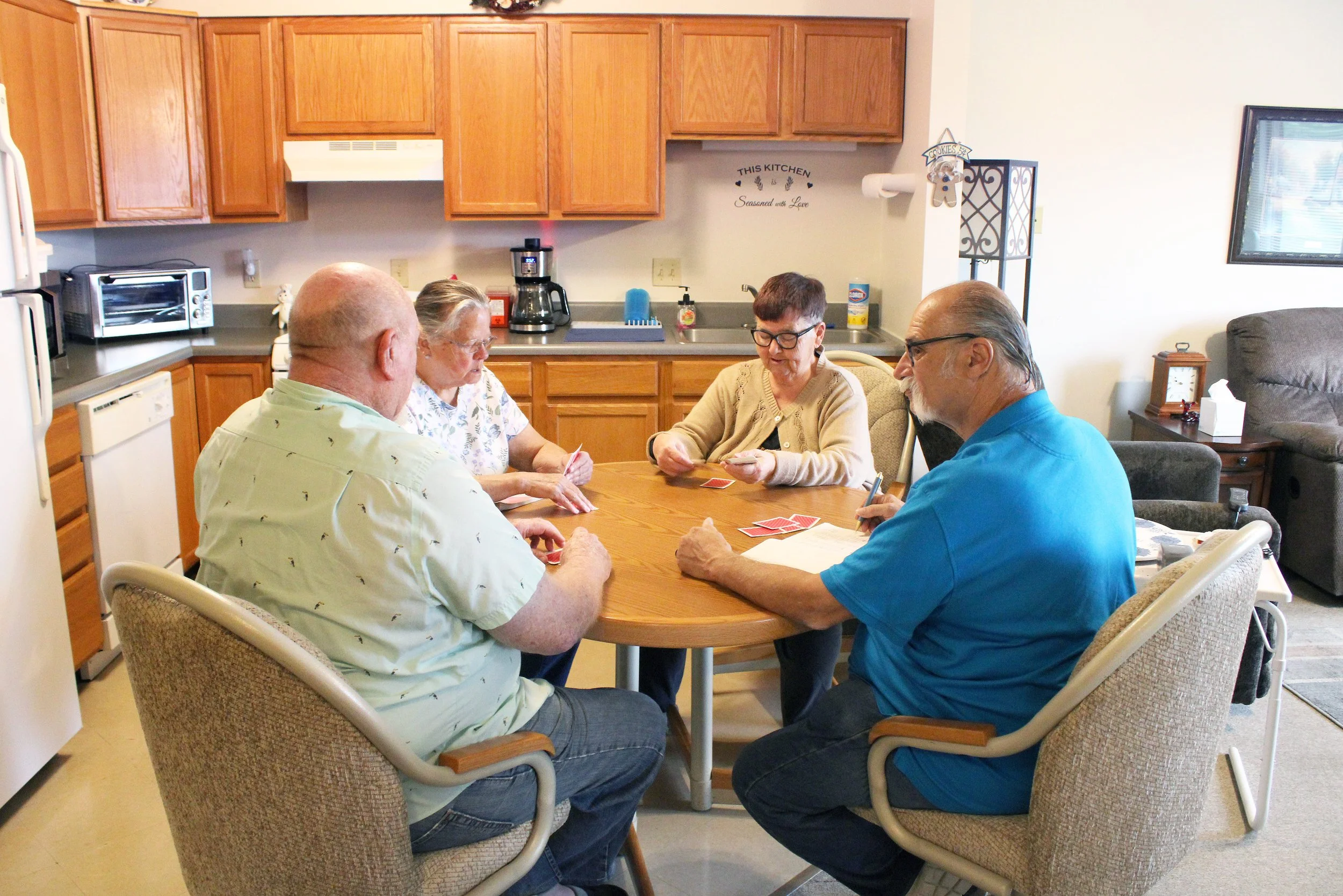 Four elderly people playing card game around a dining table in a kitchen and living room area.