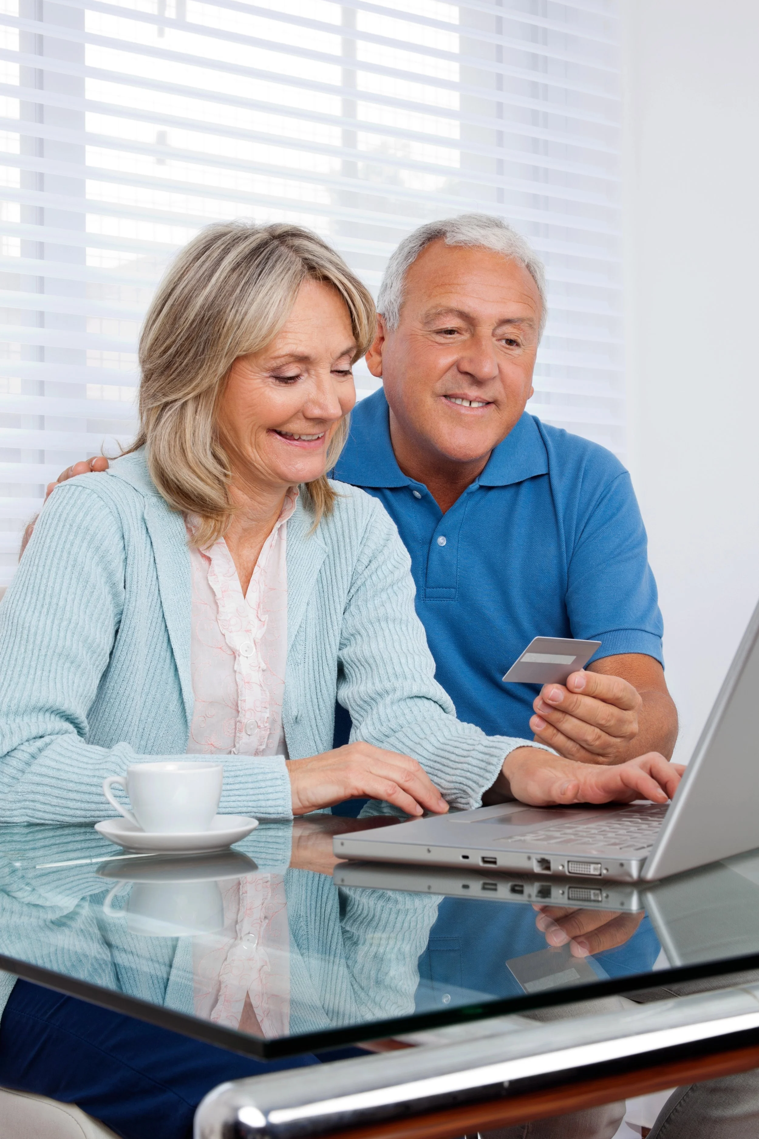 An elderly couple sitting at a table with a laptop, smiling at a smartphone, with a coffee mug on the table. They are in a bright room with white blinds.