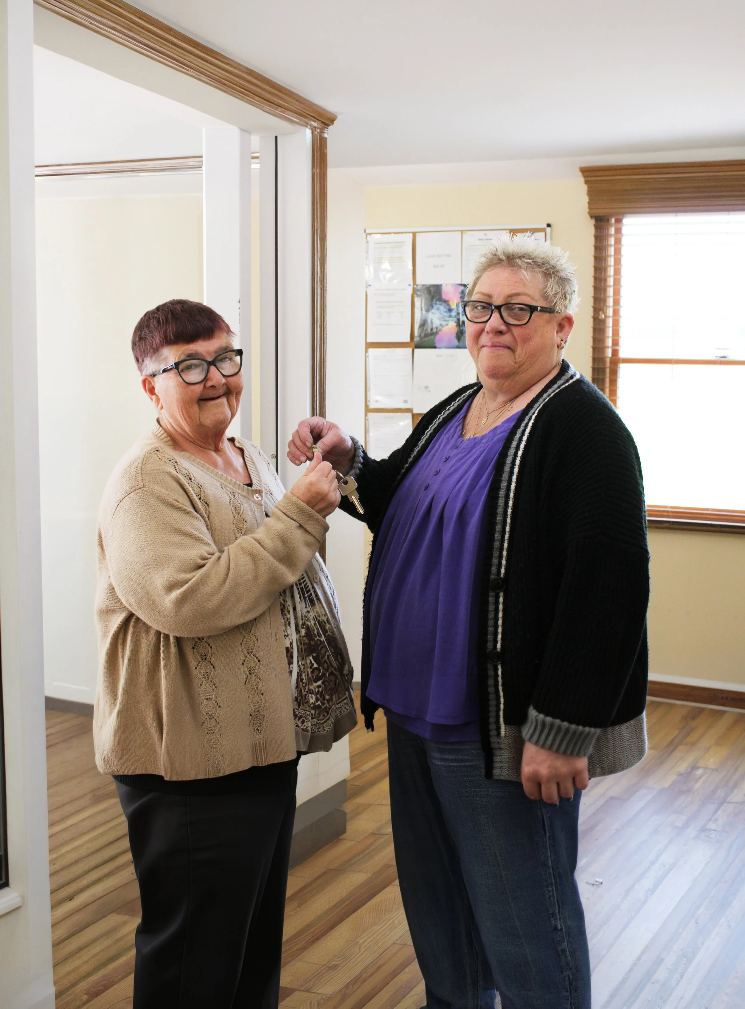 Two women, one older and one middle-aged, standing indoors in a room with wooden floors and blinds on the window. They are exchanging keys and smiling at the camera.