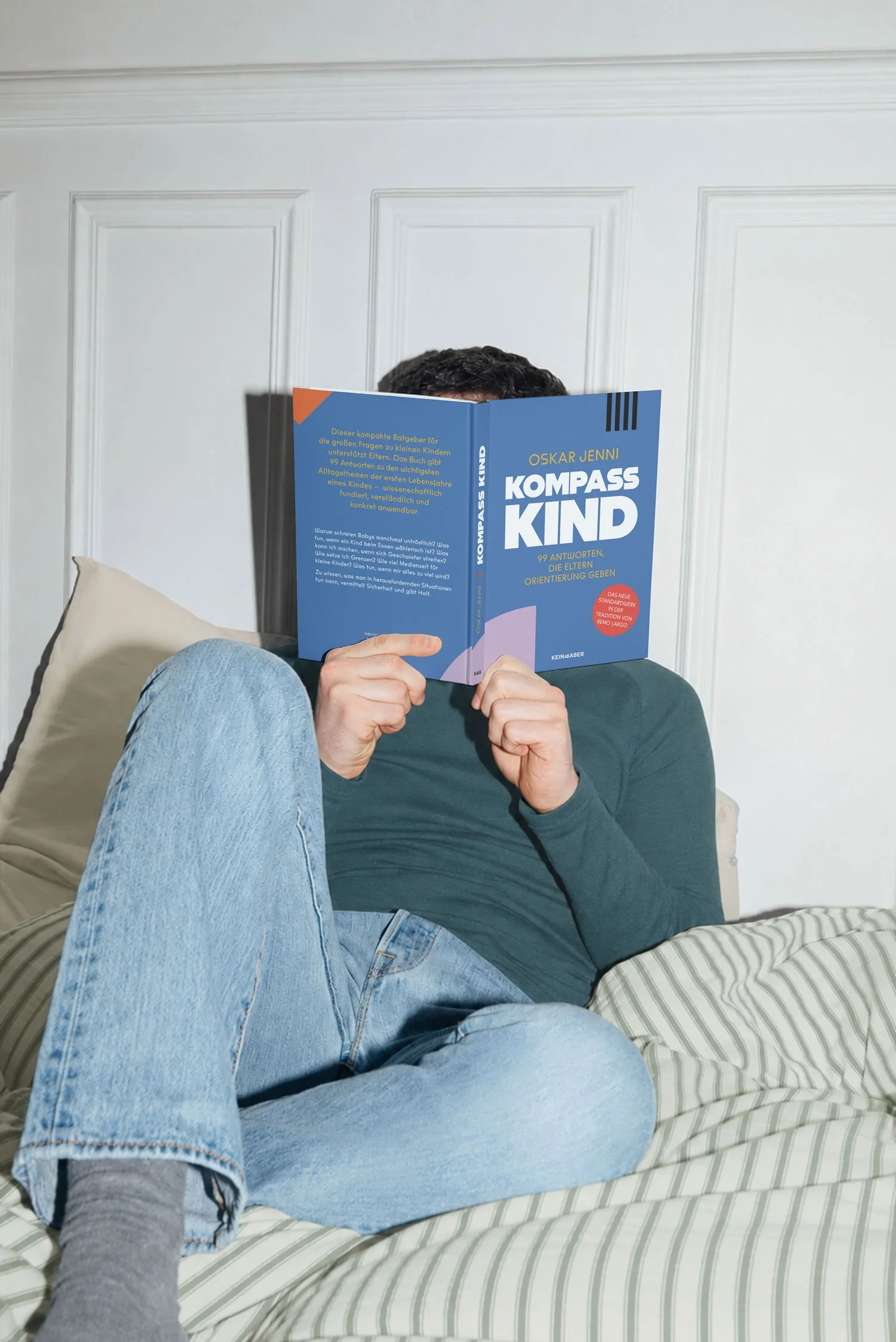 Mann sitzt entspannt auf gestreifter Bettwäsche und liest das Buch 'Kompass Kind' von Oskar Jenni – Ratgeber mit 99 Antworten für Eltern zu Erziehungsfragen