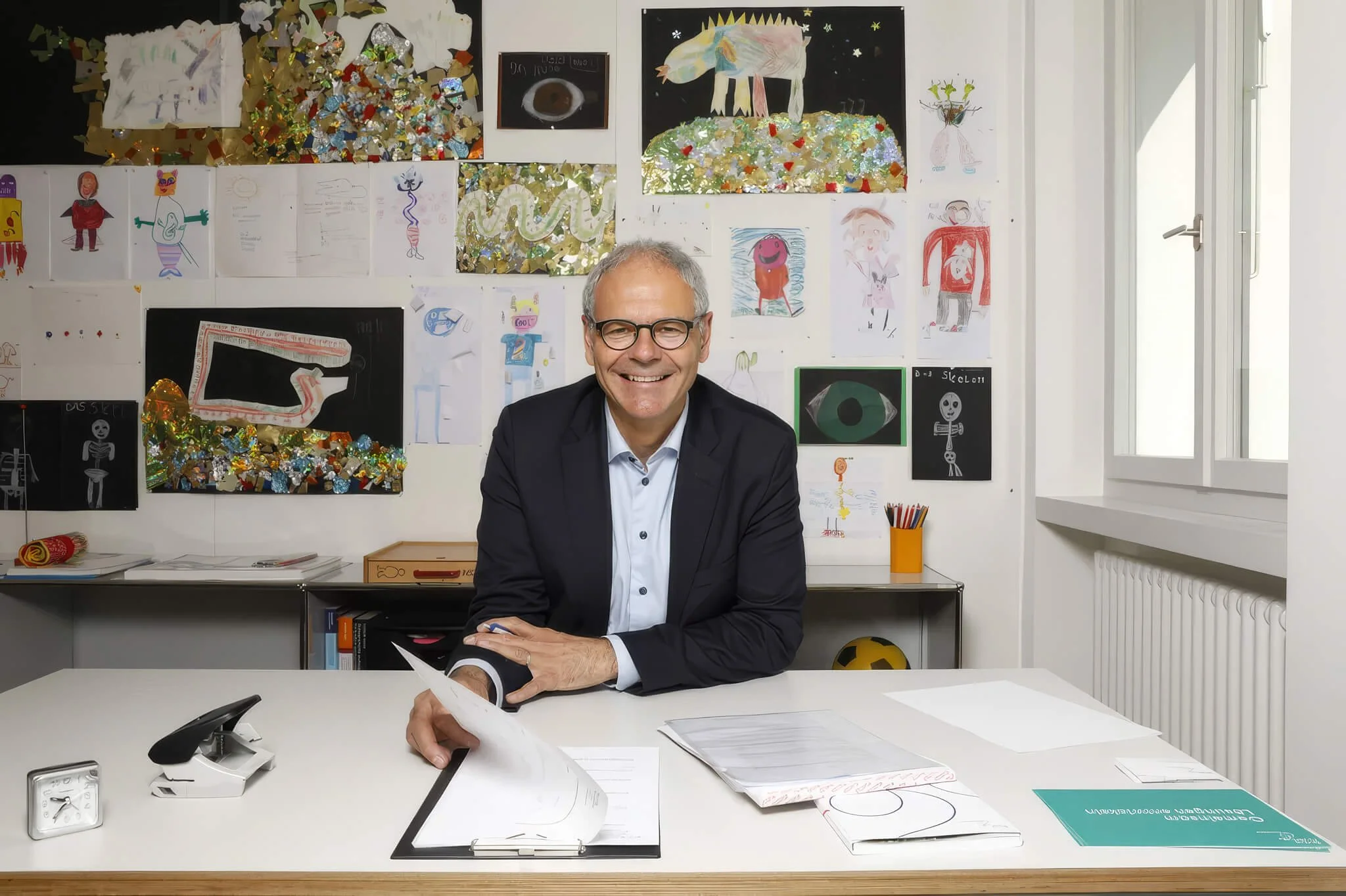 Porträtfoto von Oskar Jenni, lächelnd im dunklen Sakko an einem Schreibtisch sitzend, im Hintergrund eine Wand mit bunten Kinderzeichnungen, im Vordergrund liegen Unterlagen auf dem Tisch.