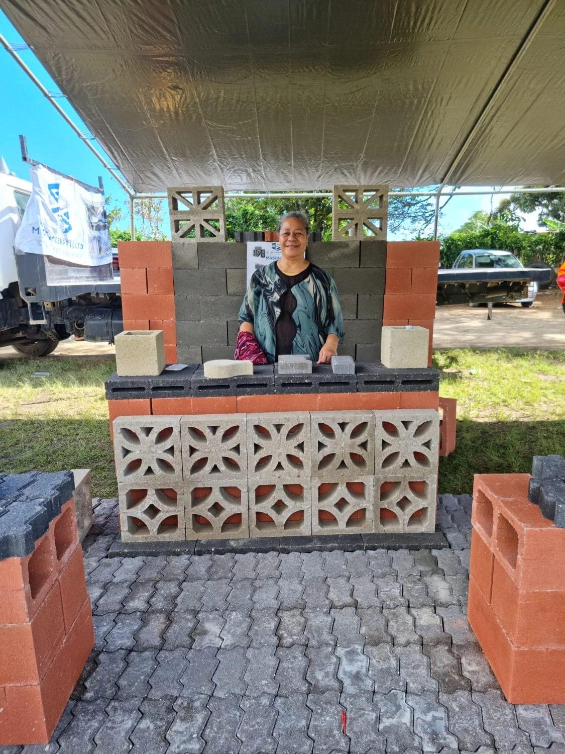 A woman standing behind a brick and cinder block display at an outdoor event under a canopy, with trees and vehicles in the background.