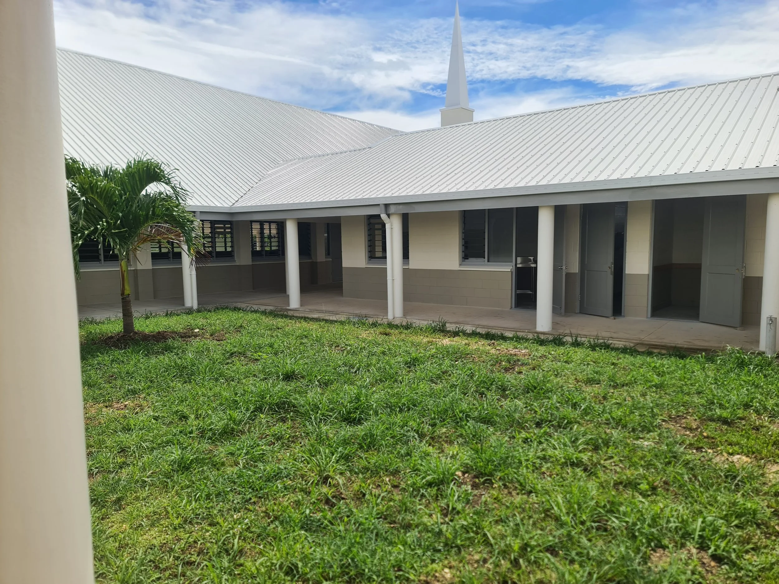 A building with a white metal roof, surrounded by a grassy yard, with a palm tree in the foreground and a partly cloudy blue sky in the background.