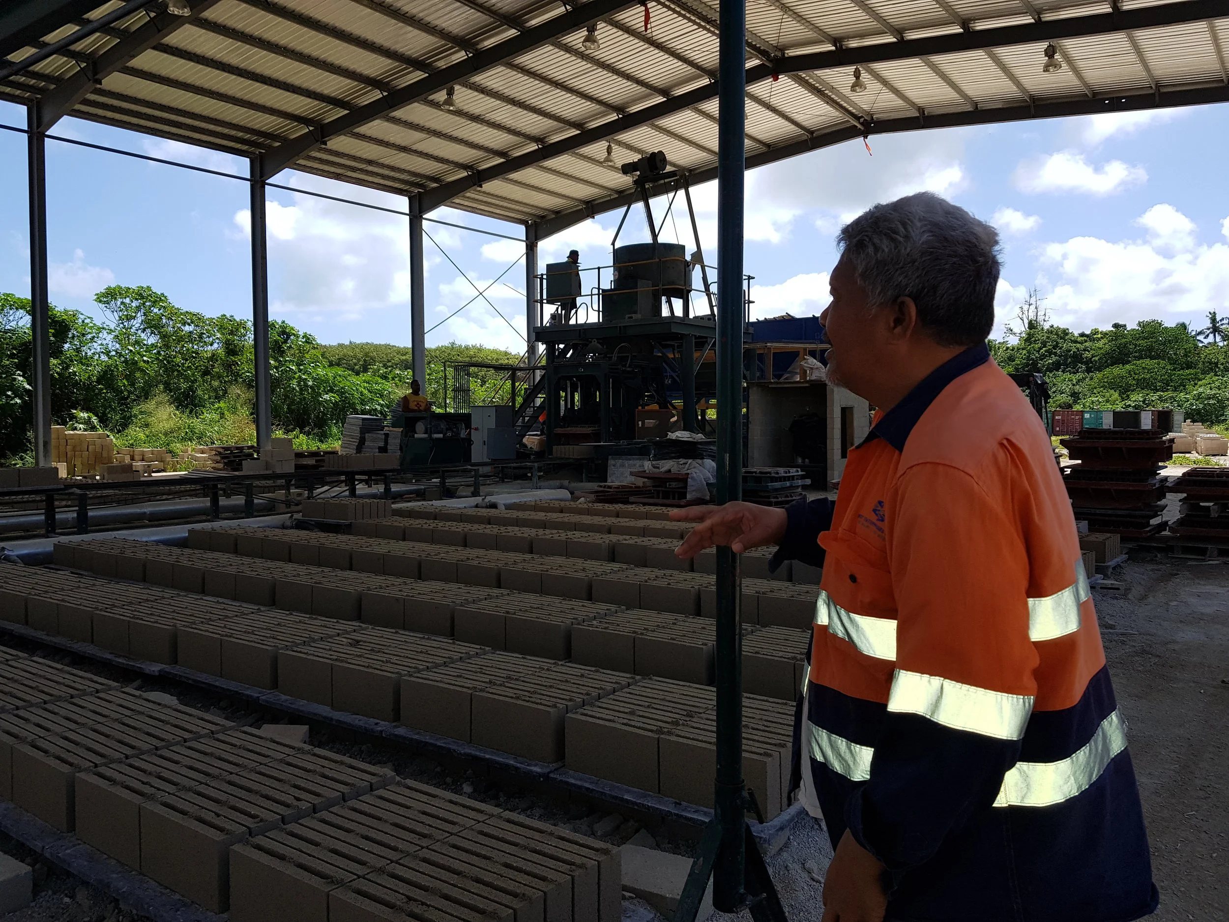 Workers on a construction site under a metal roof, with stacks of bricks and greenery outside.