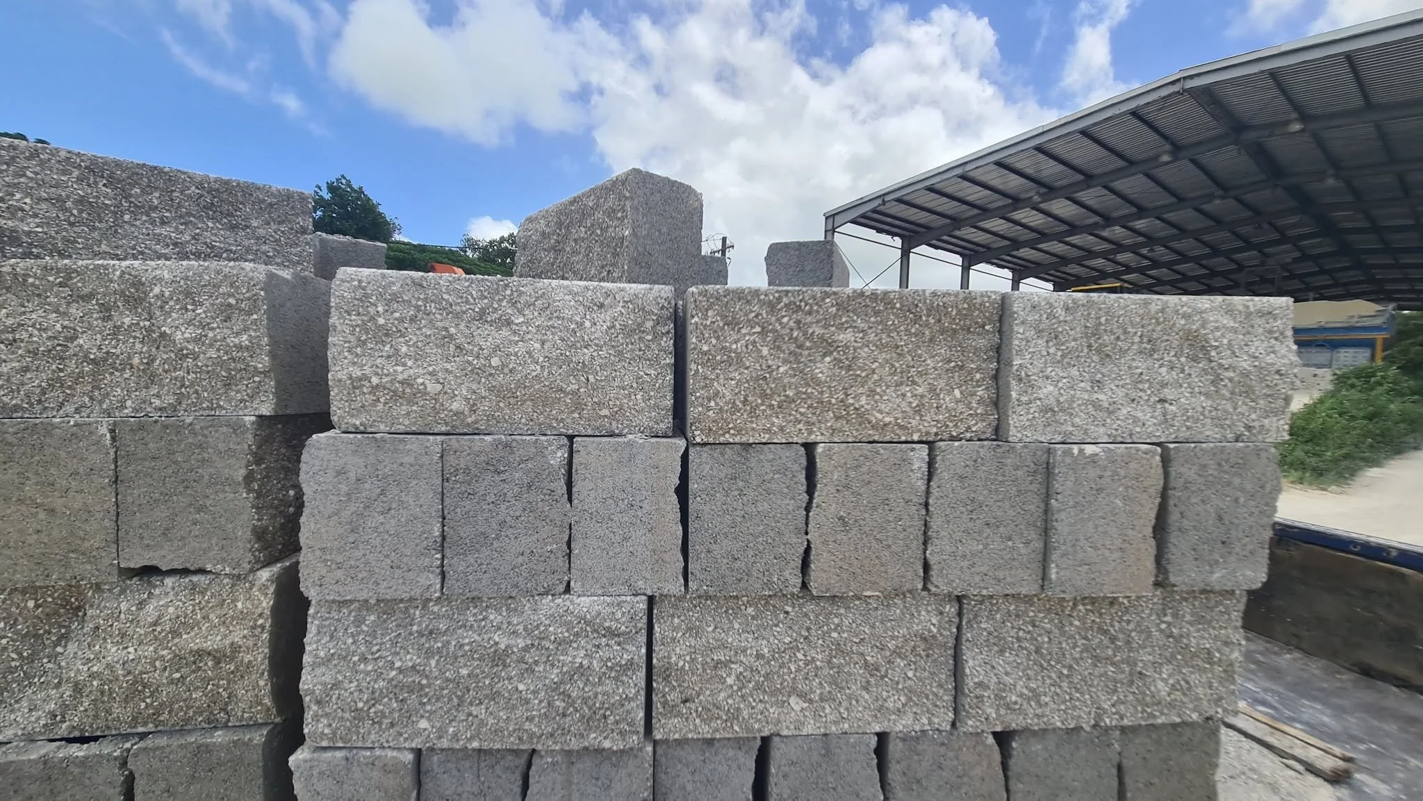 Stacked concrete blocks in an outdoor storage area with a partially covered structure and a partly cloudy sky.