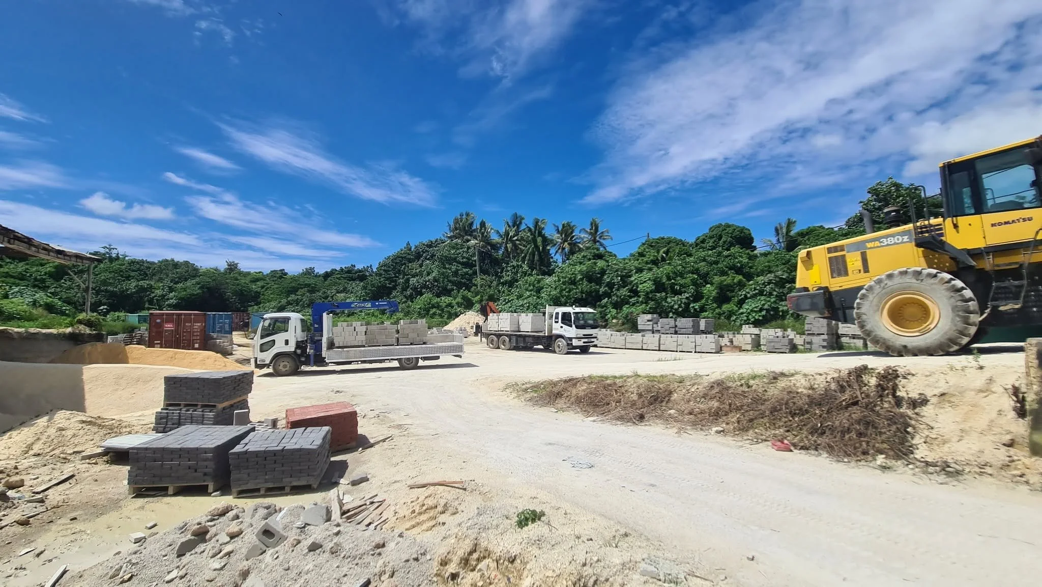 Construction site with trucks, construction equipment, and building materials against a blue sky with scattered clouds.