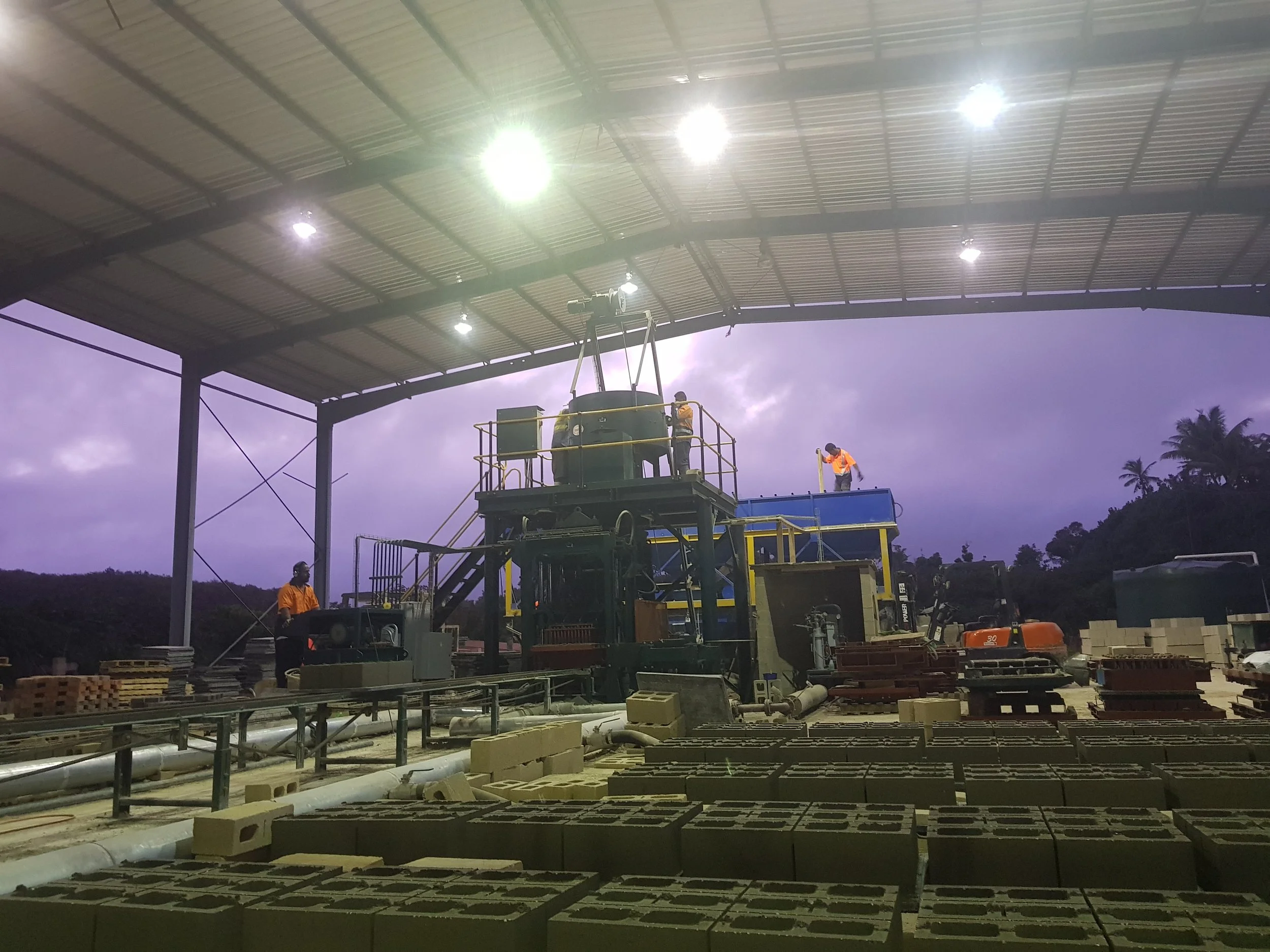 Construction workers installing lighting under a large roof structure during dusk or early evening.