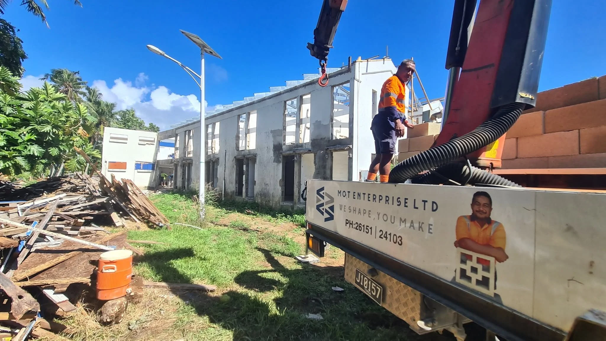 Construction site with workers building a white structure, blue sky, and green trees, with debris and construction materials on the ground, and a truck visible in the foreground.