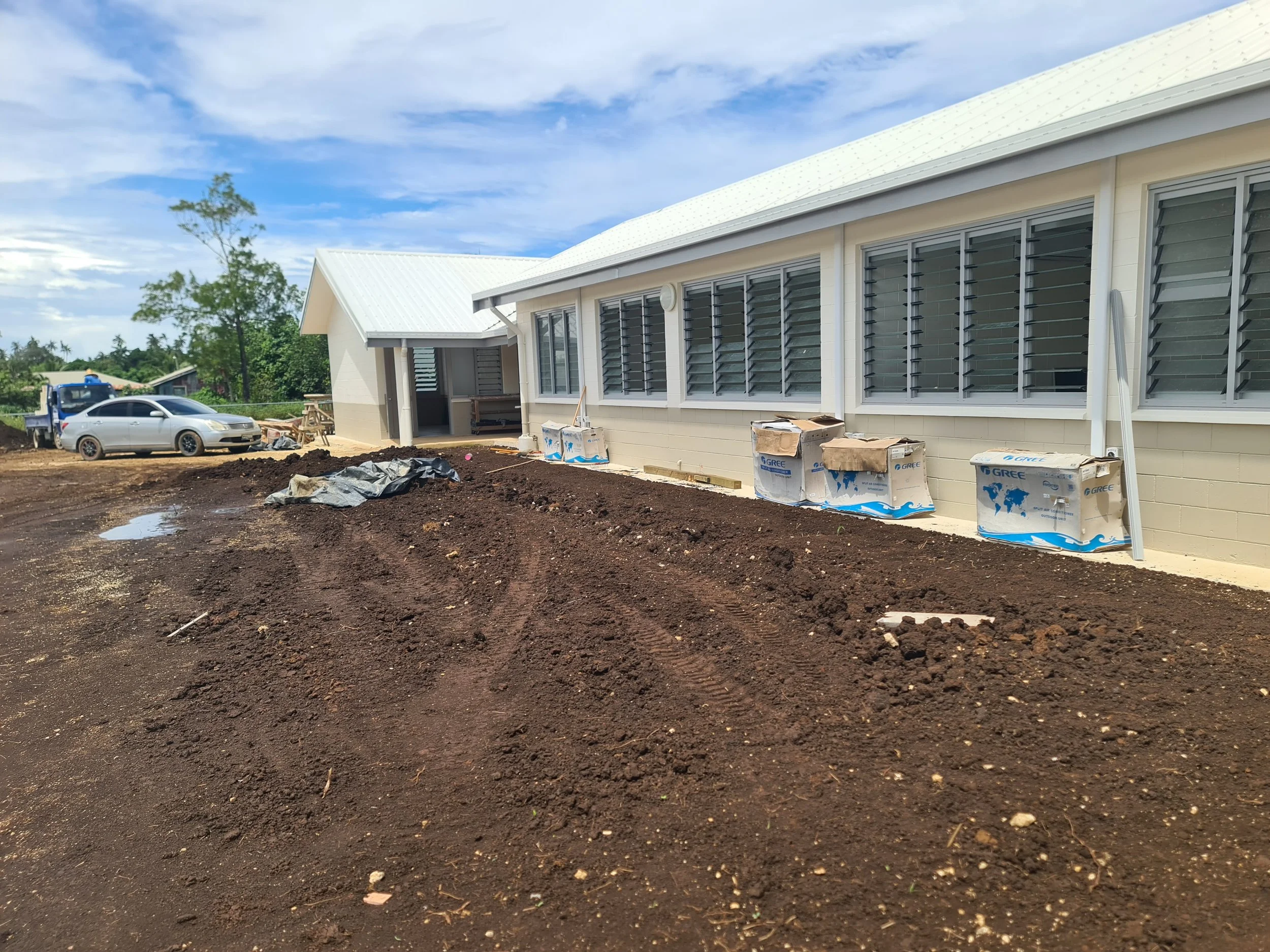 Newly built house with white exterior, large windows with shutters, and a metal roof. Construction materials and vehicles are present on the dirt yard, indicating ongoing construction.