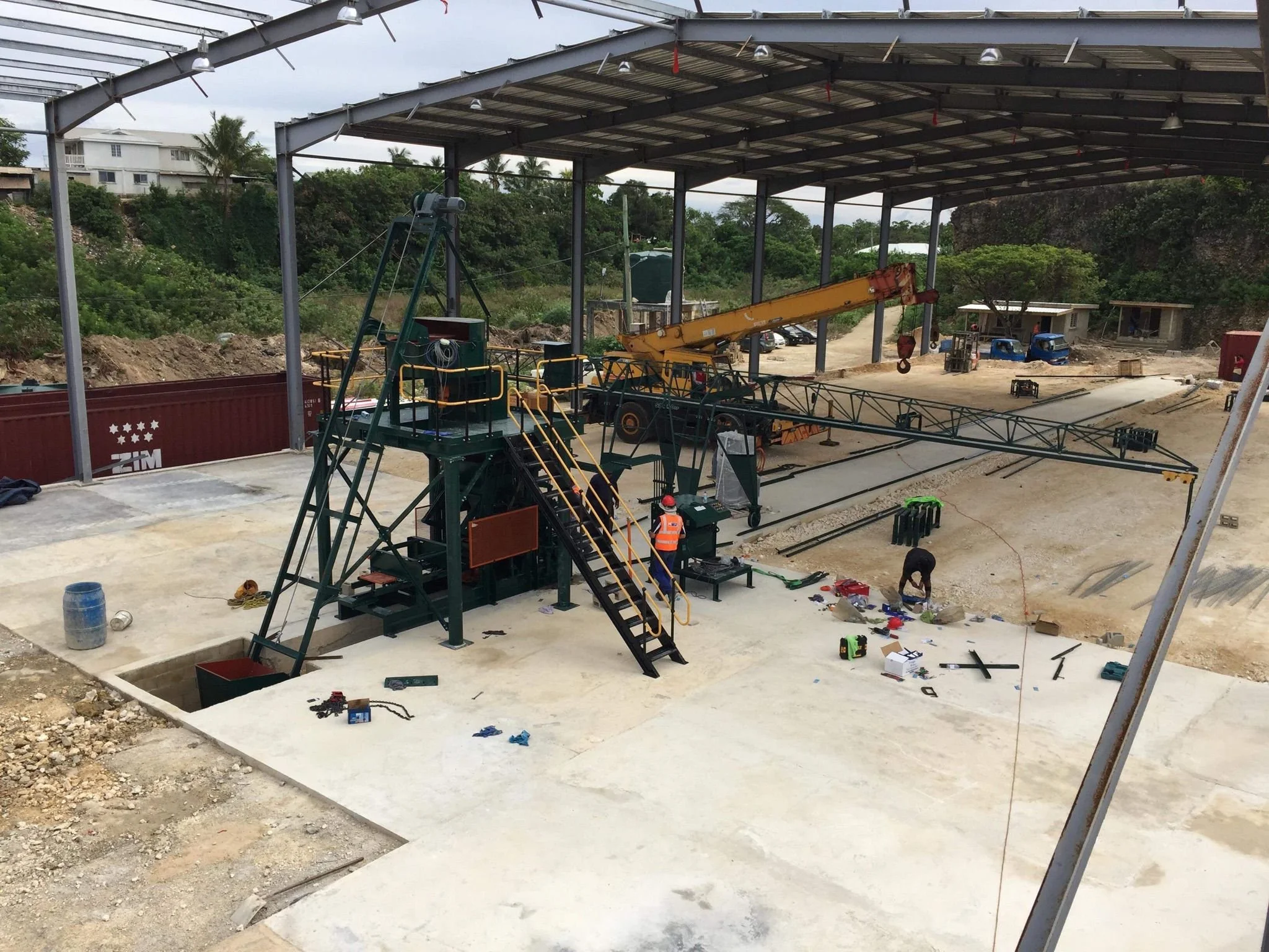 Construction site with a steel framework for a roof, construction equipment, and workers. Partially completed concrete floor, dirt, and scattered tools.