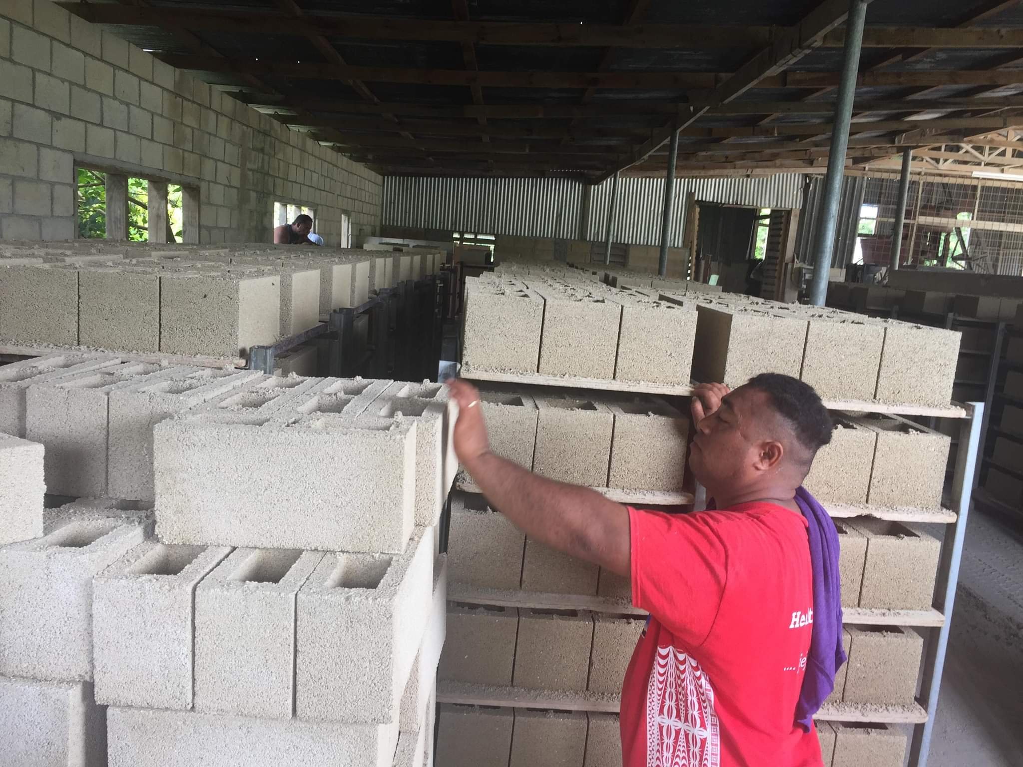 People working on constructing a building with concrete blocks and metal supports, inside a partially built structure with a wooden roof.