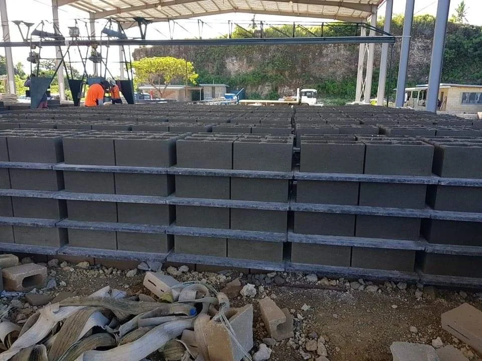 Stacked concrete blocks at a construction site with workers in the background under a covered structure.